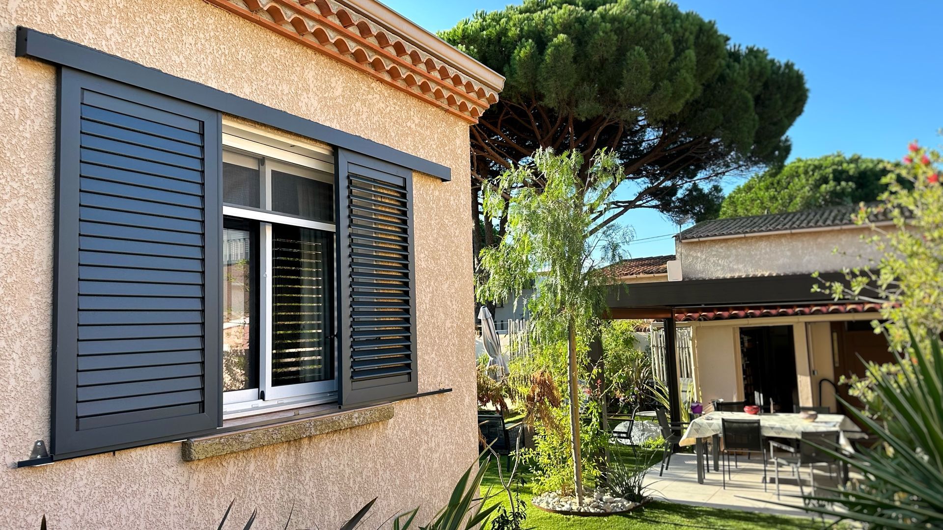 Maison avec des volets noirs sur une fenêtre, donnant sur une cour arrière avec un patio et une deuxième maison.