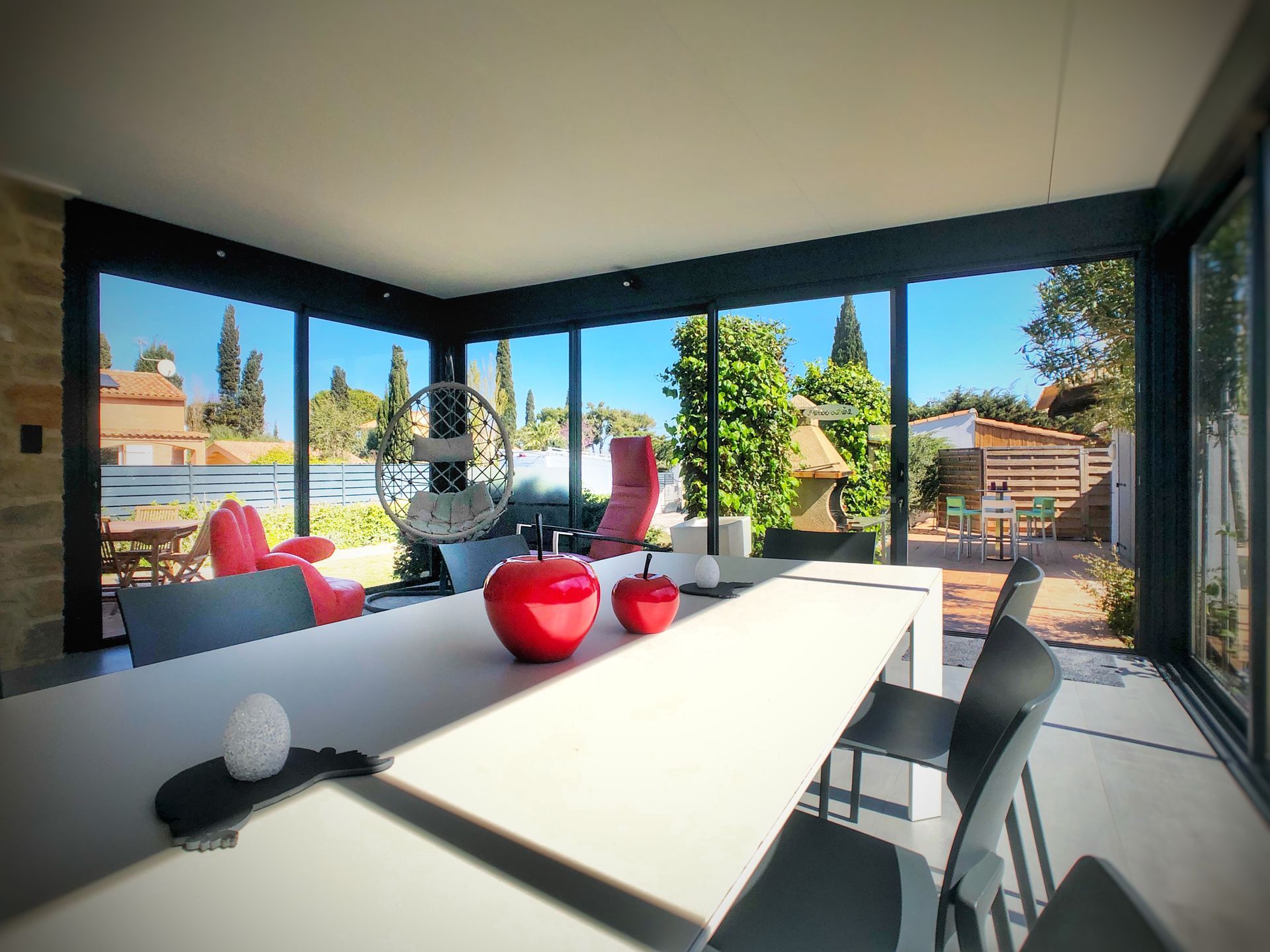 Salle à manger avec de grandes fenêtres donnant sur une cour, une longue table et des pommes rouges décoratives.
