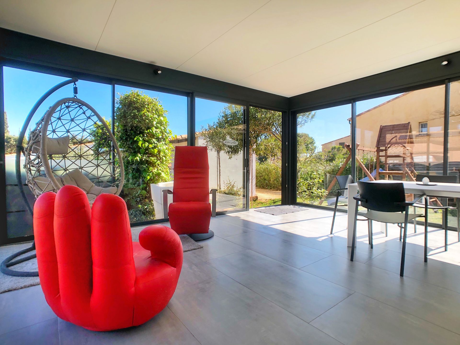 Véranda avec de grandes fenêtres donnant sur une cour arrière ; une chaise suspendue et une table avec des chaises.