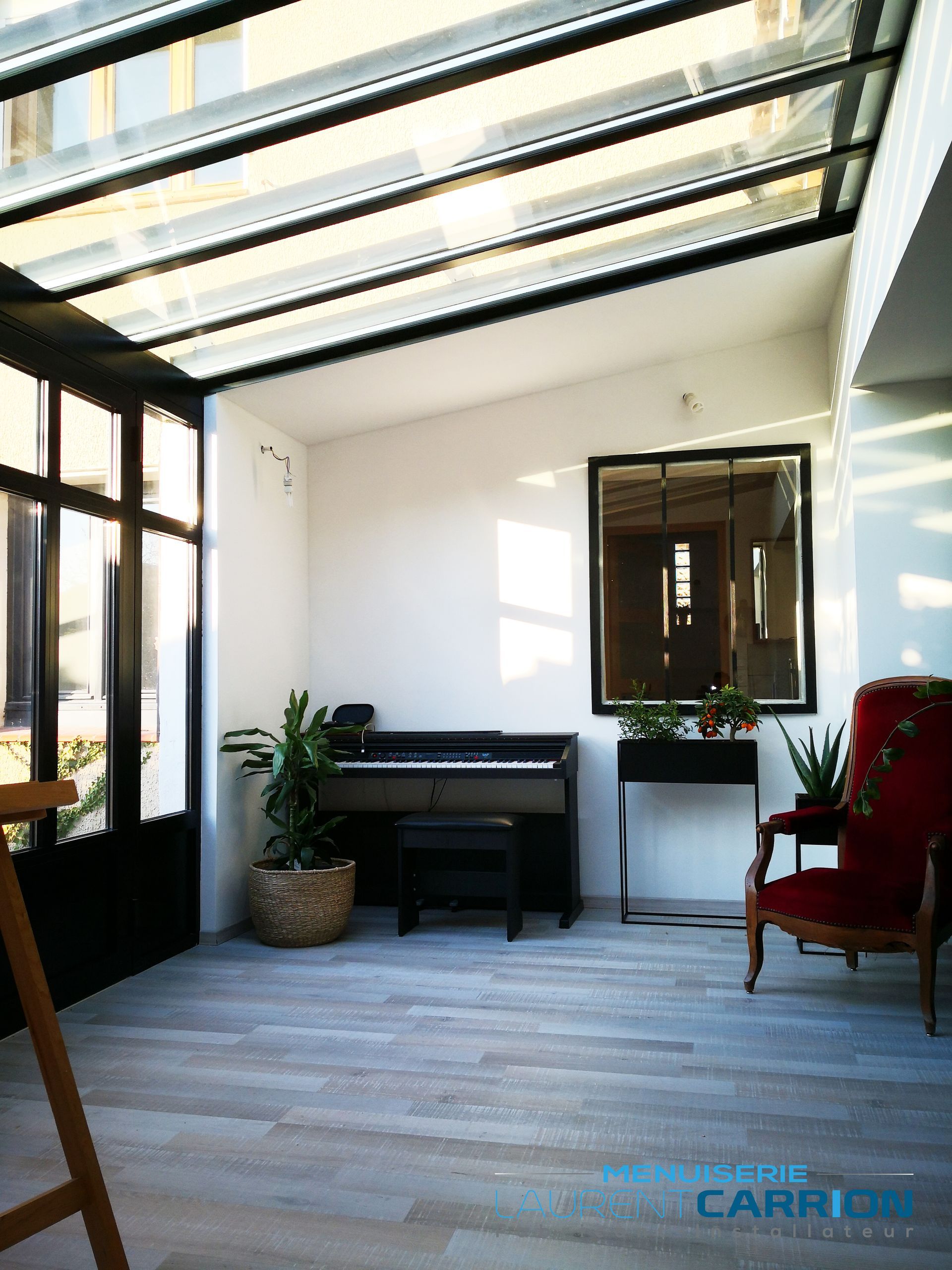 Véranda avec toit en verre, piano, chaise rouge et plantes ; sol gris clair et murs blancs.