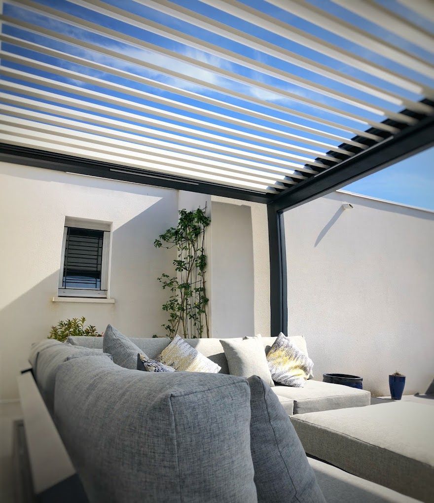 Terrasse extérieure avec canapé blanc, coussins et persiennes réglables sous un ciel lumineux.