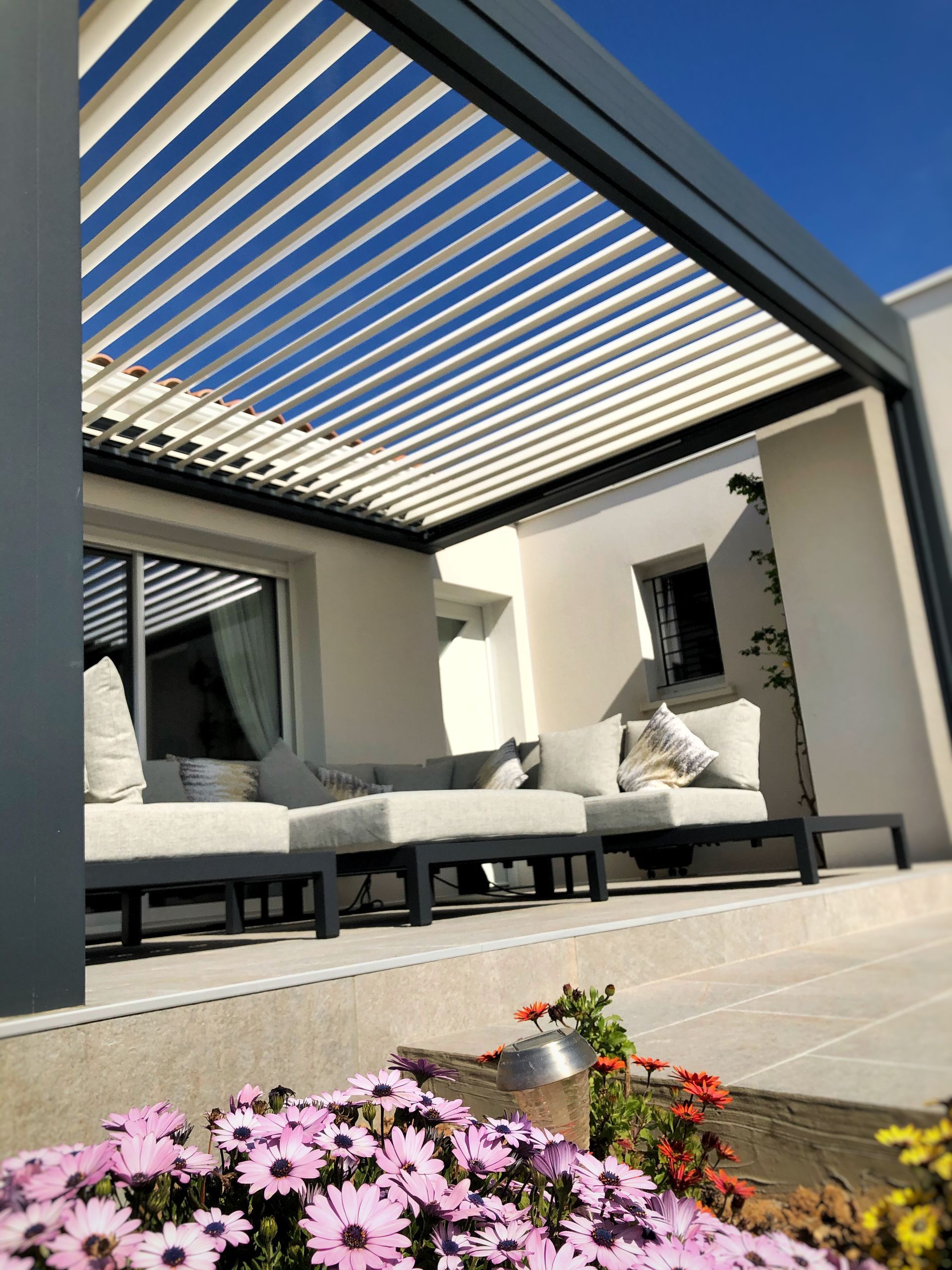 Terrasse extérieure avec mobilier gris, pergola et fleurs.