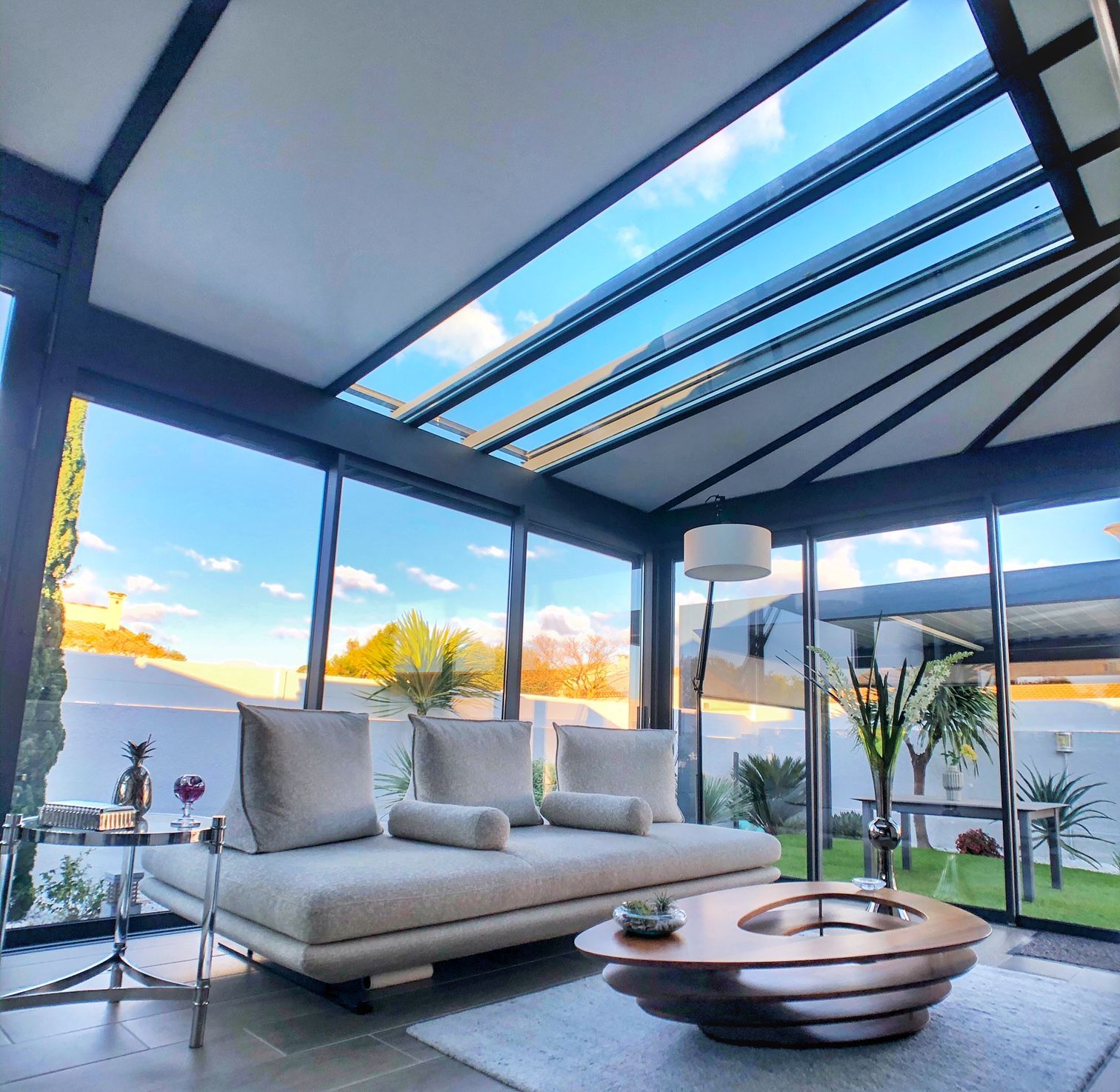 Véranda avec murs et plafond en verre, meublée d'un canapé et d'une table basse, avec vue sur le ciel et la cour.