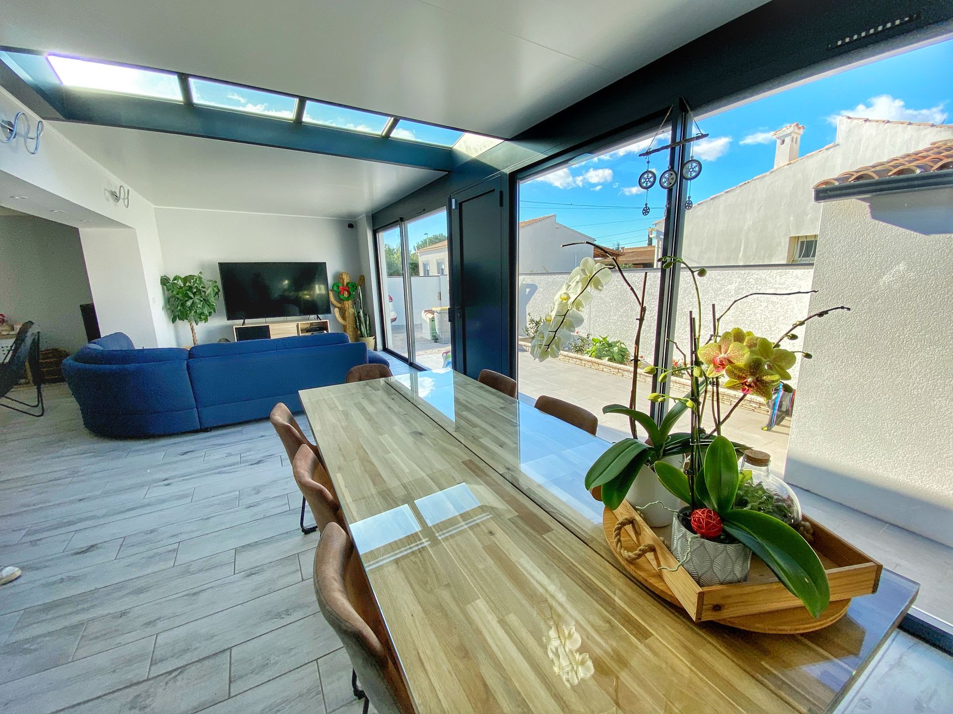 Salle à manger moderne avec table en bois, canapé bleu, grandes fenêtres et plantes ; vue extérieure ensoleillée.