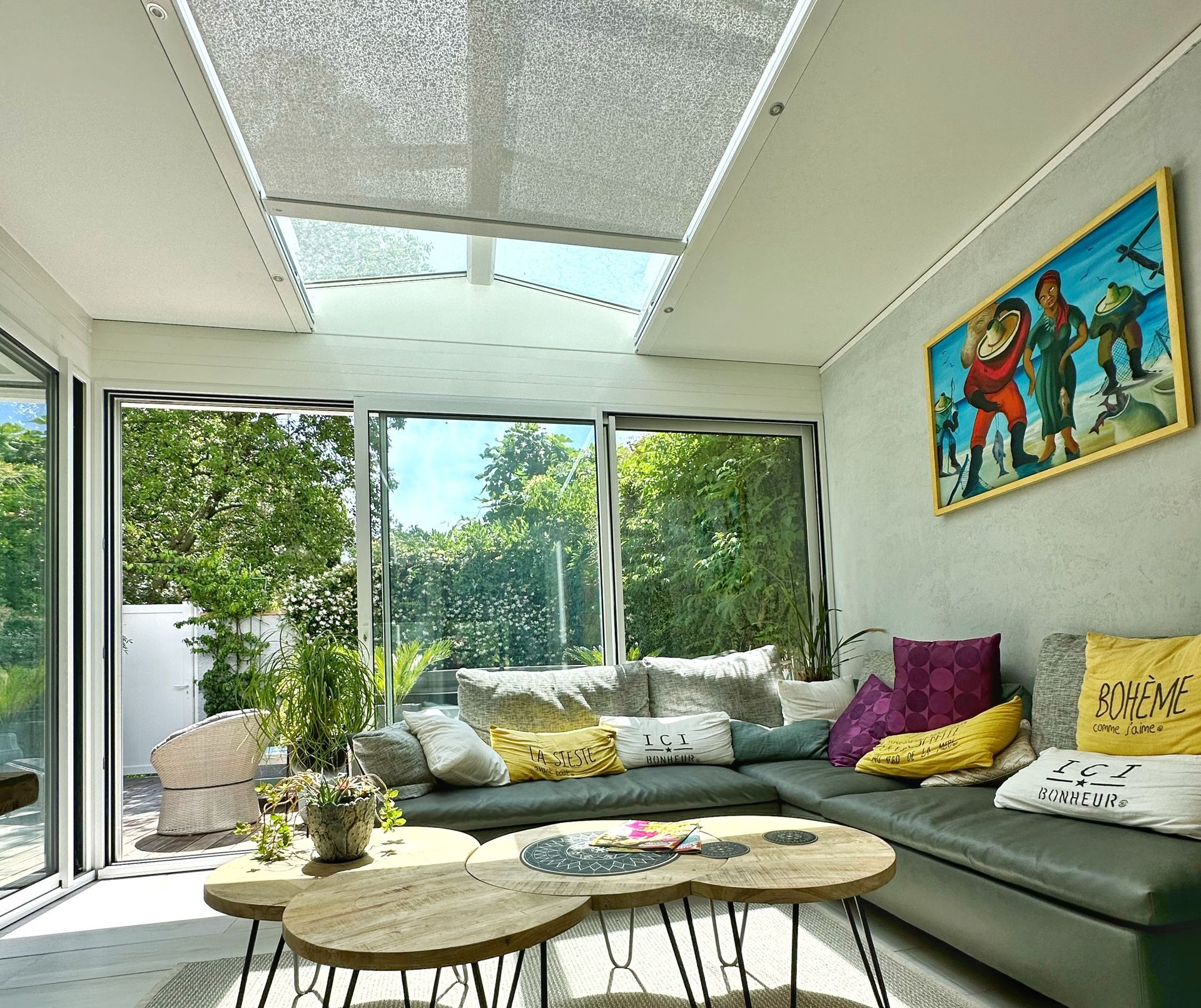 Véranda avec section grise, tables basses en bois, coussins colorés et puits de lumière.