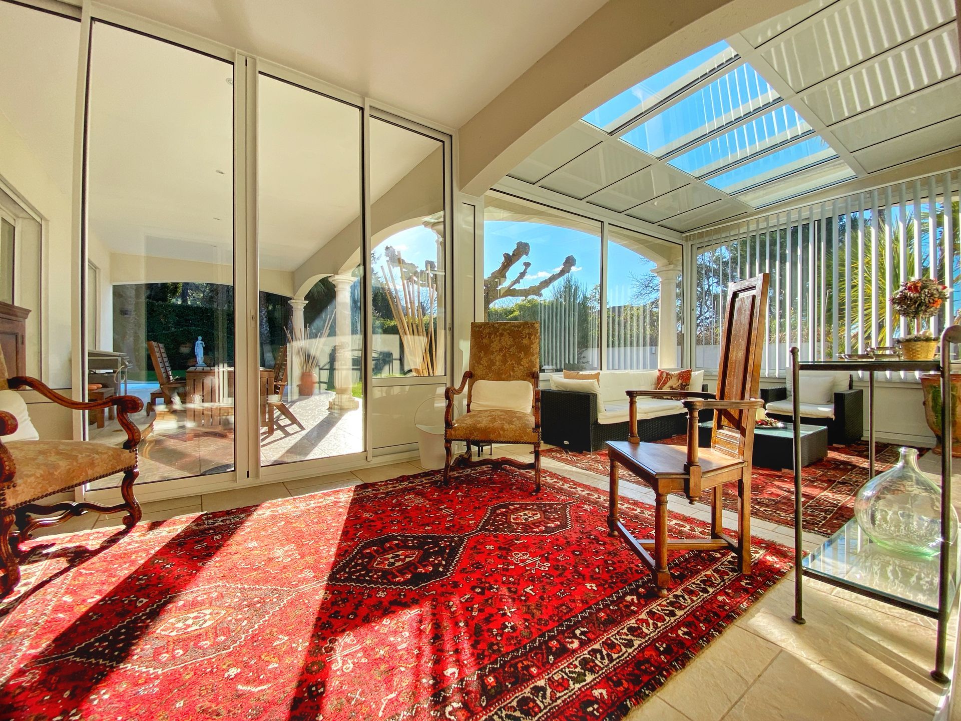 Véranda avec tapis rouge, parois vitrées et meubles anciens. Vue extérieure ensoleillée.