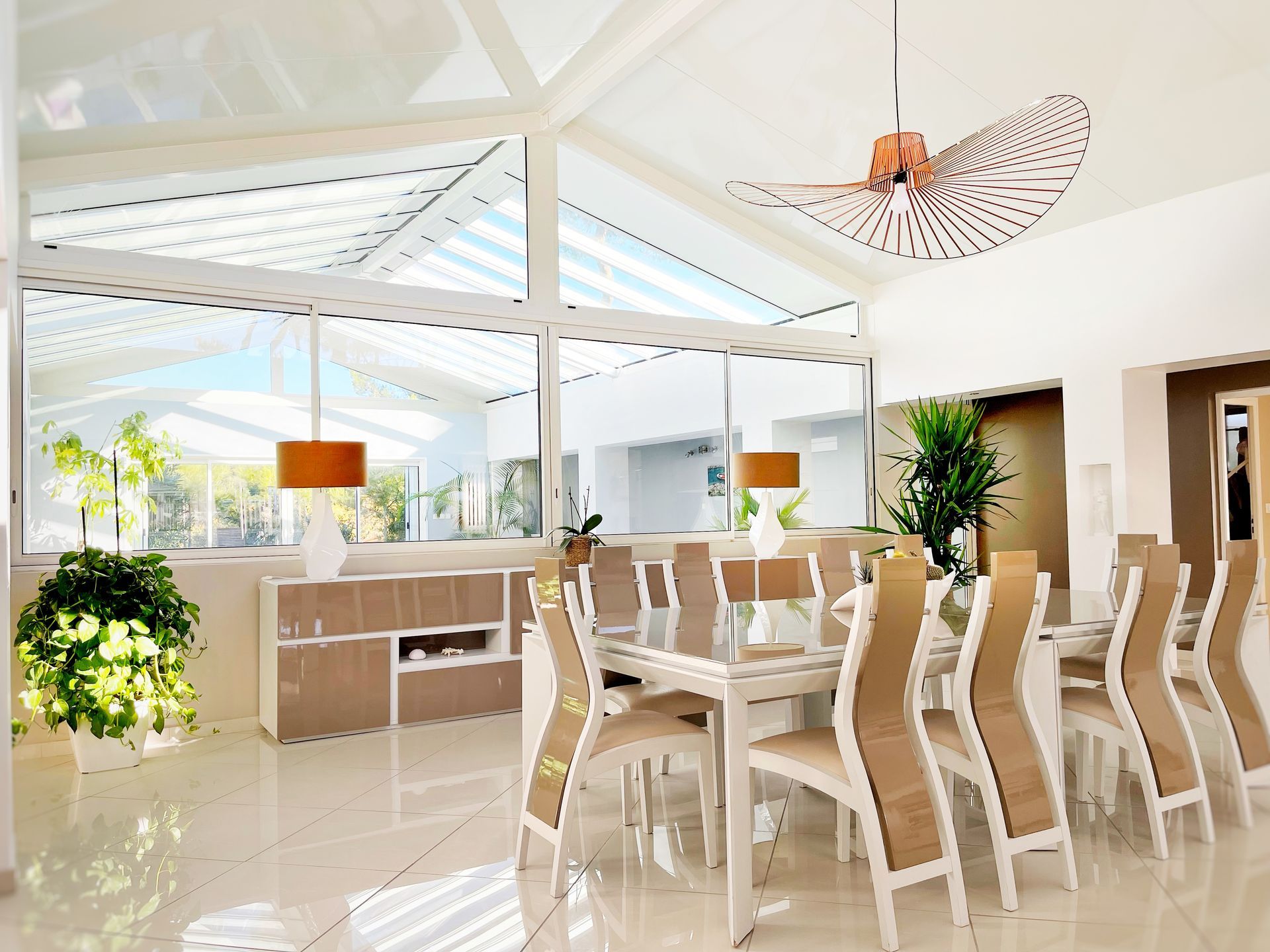 Salle à manger lumineuse avec de grandes fenêtres, des meubles blancs et beiges et un luminaire moderne.