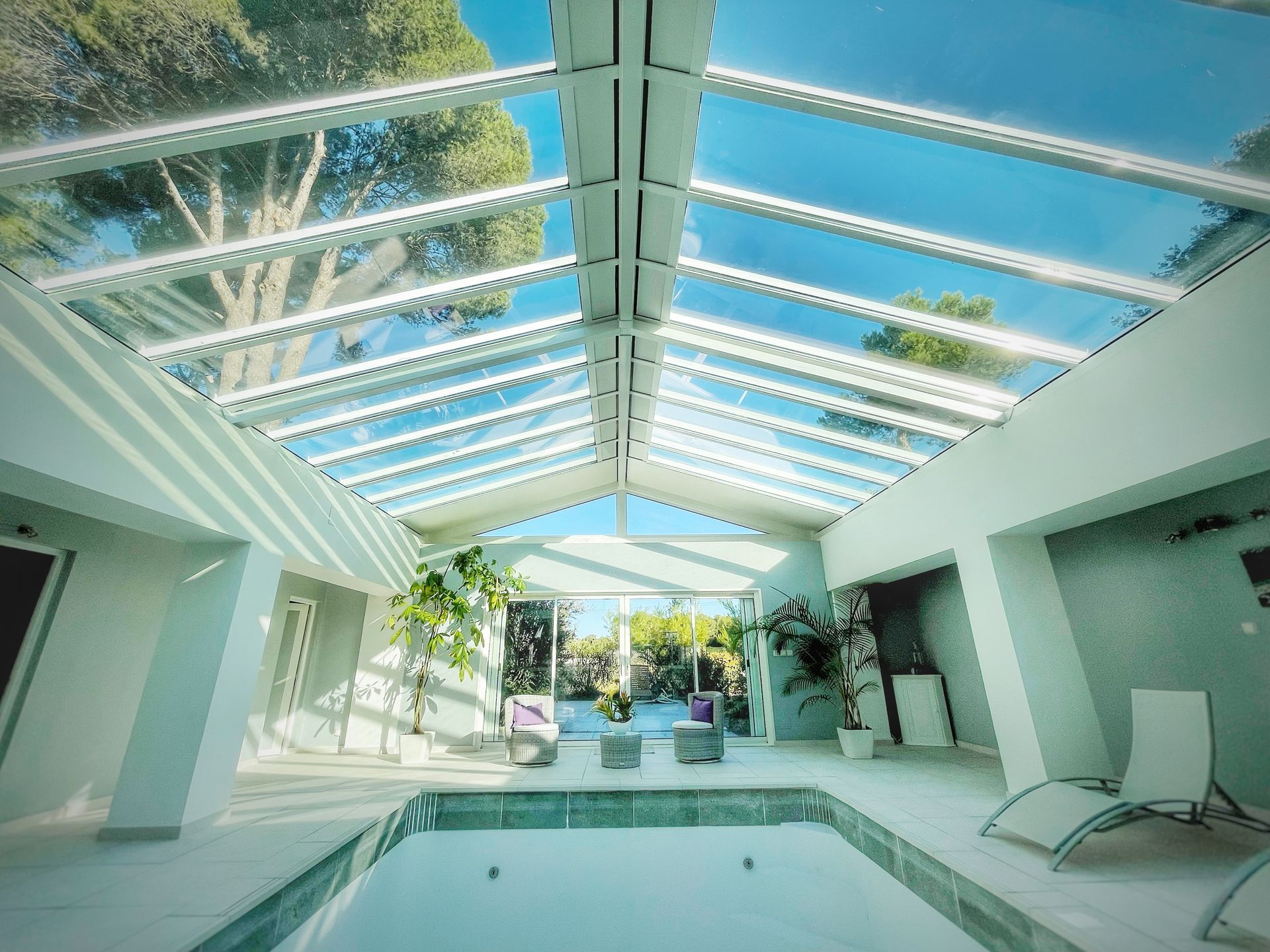 Piscine intérieure avec verrière, lumière naturelle. Murs blancs, arbres visibles, chaises longues.