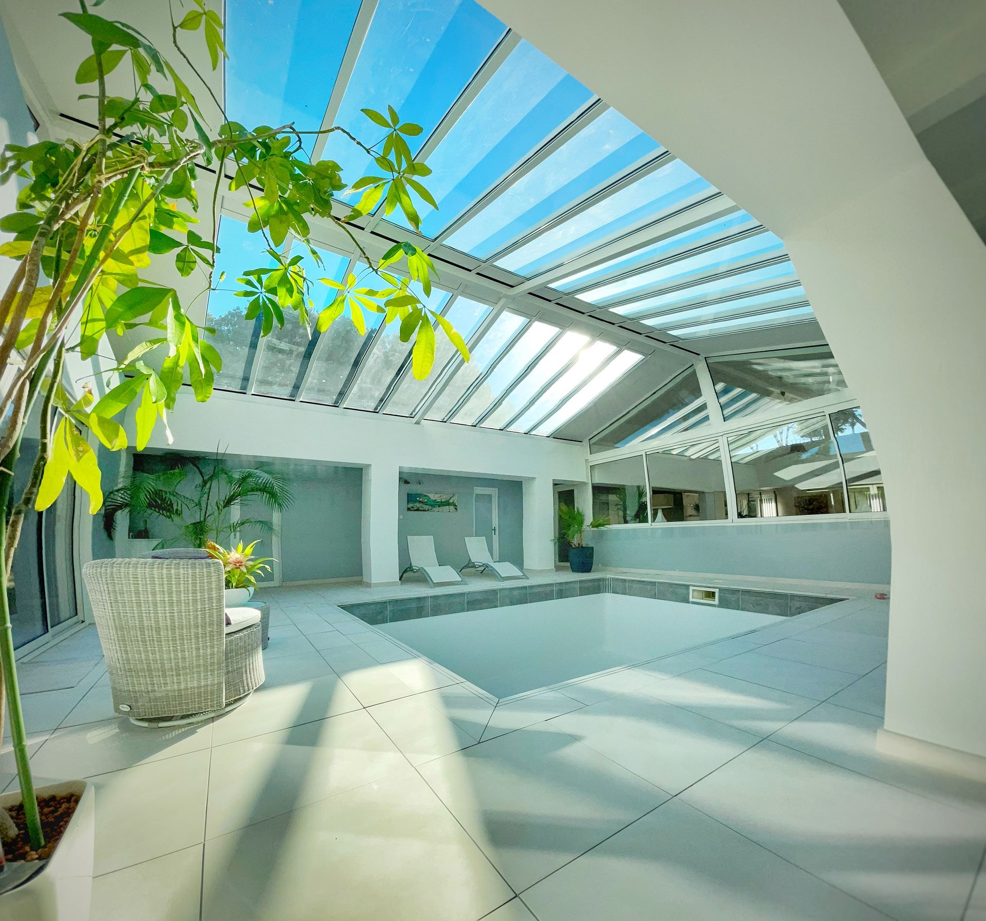 Piscine intérieure sous un toit en verre, avec chaises longues, murs blancs et lumière du soleil qui entre à flot.