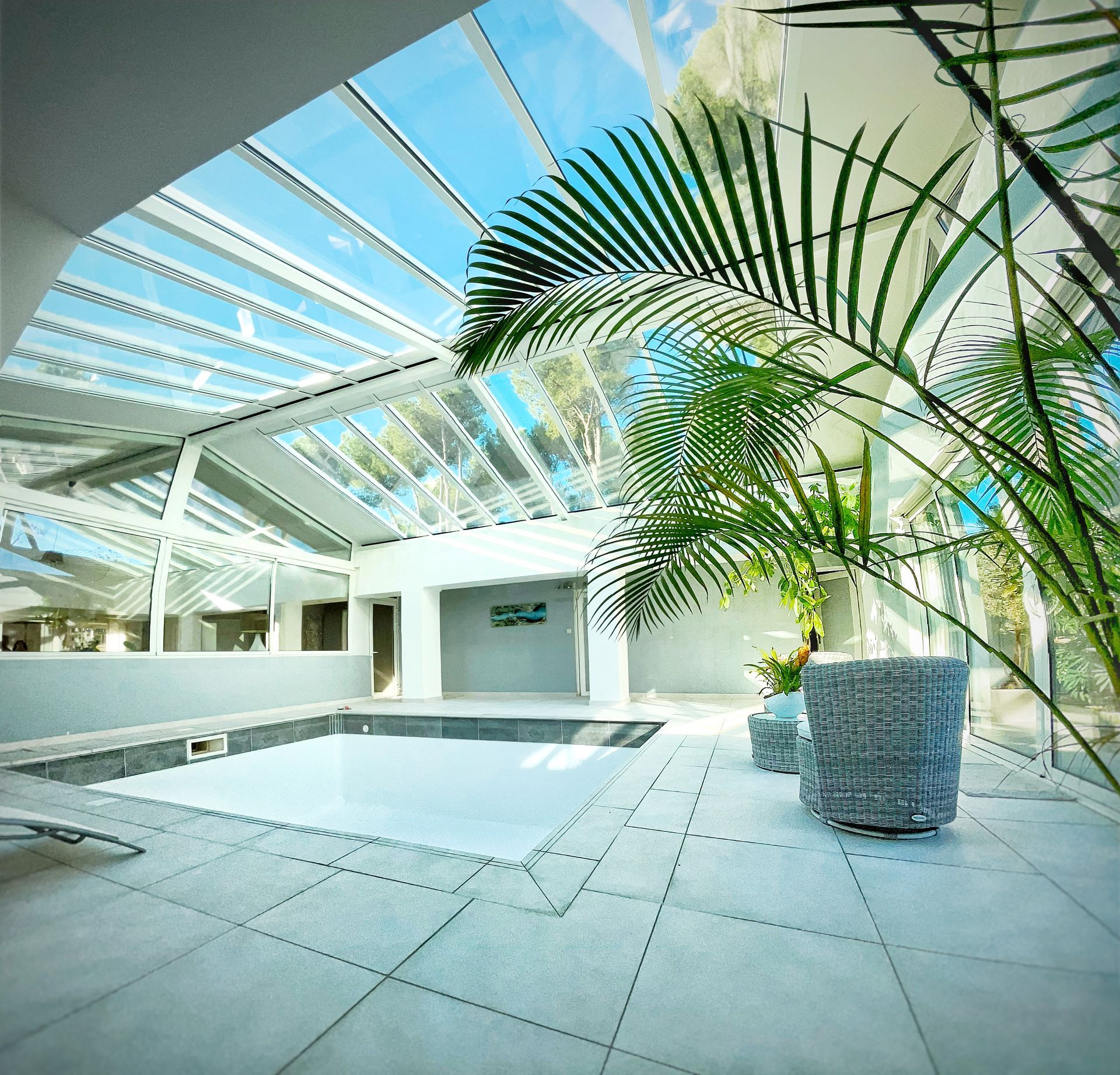 Piscine intérieure avec verrière, palmiers et jardinières en pierre. Ciel bleu éclatant visible.