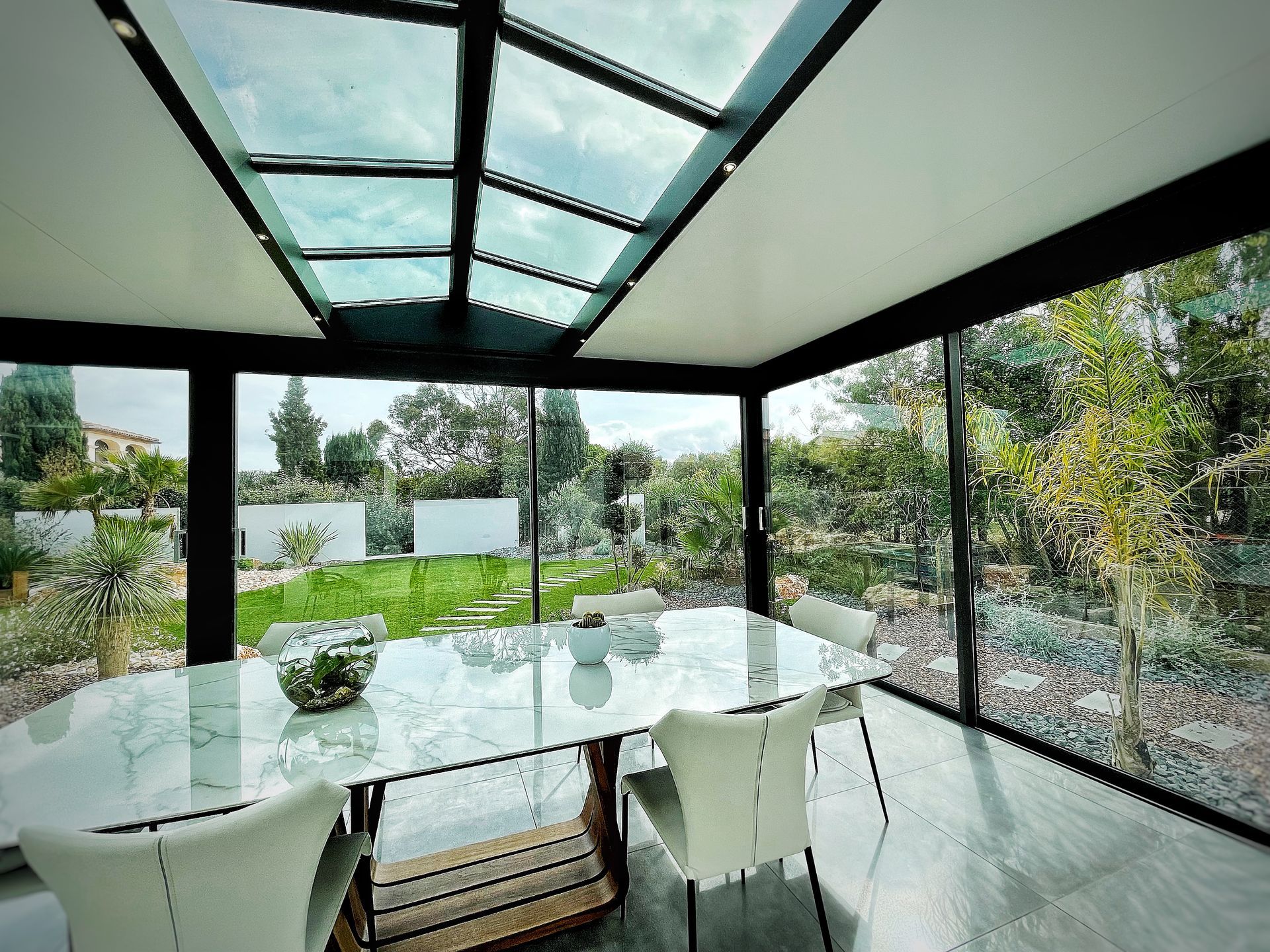 Véranda avec toit et murs en verre ; table à manger avec chaises blanches donnant sur un jardin.
