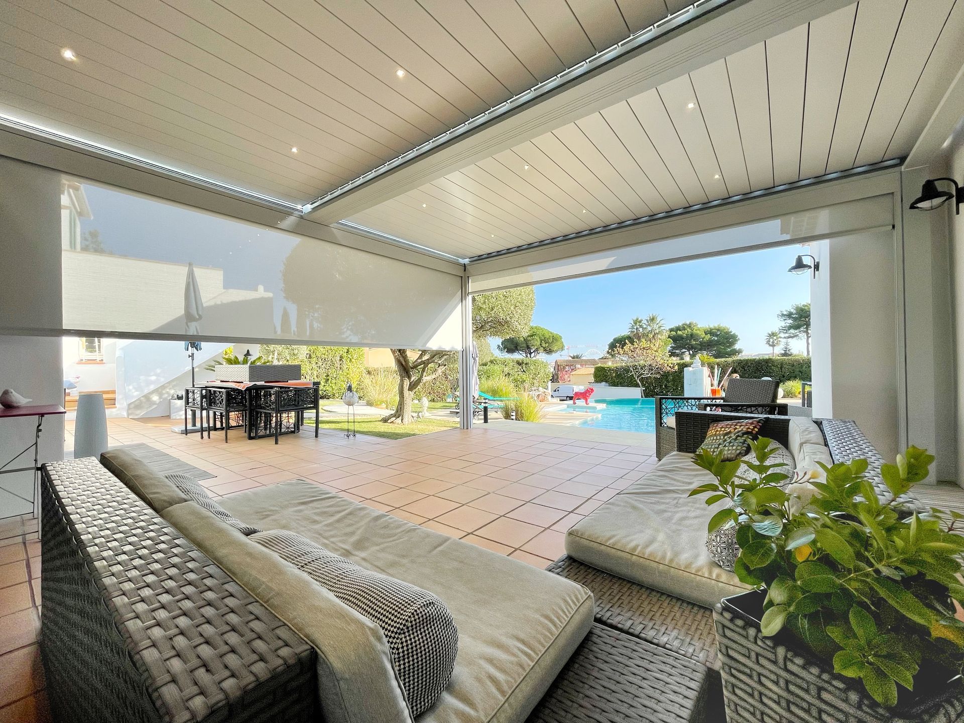 Terrasse extérieure avec toit rétractable, vue sur la piscine, sièges et plante en pot.