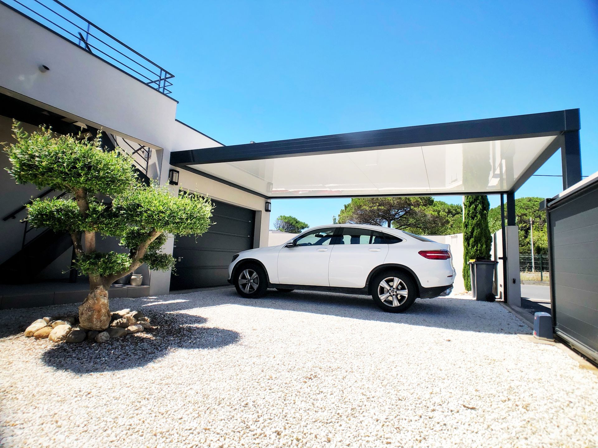 Voiture blanche sous un abri d'auto à cadre noir devant une maison moderne par une journée ensoleillée.