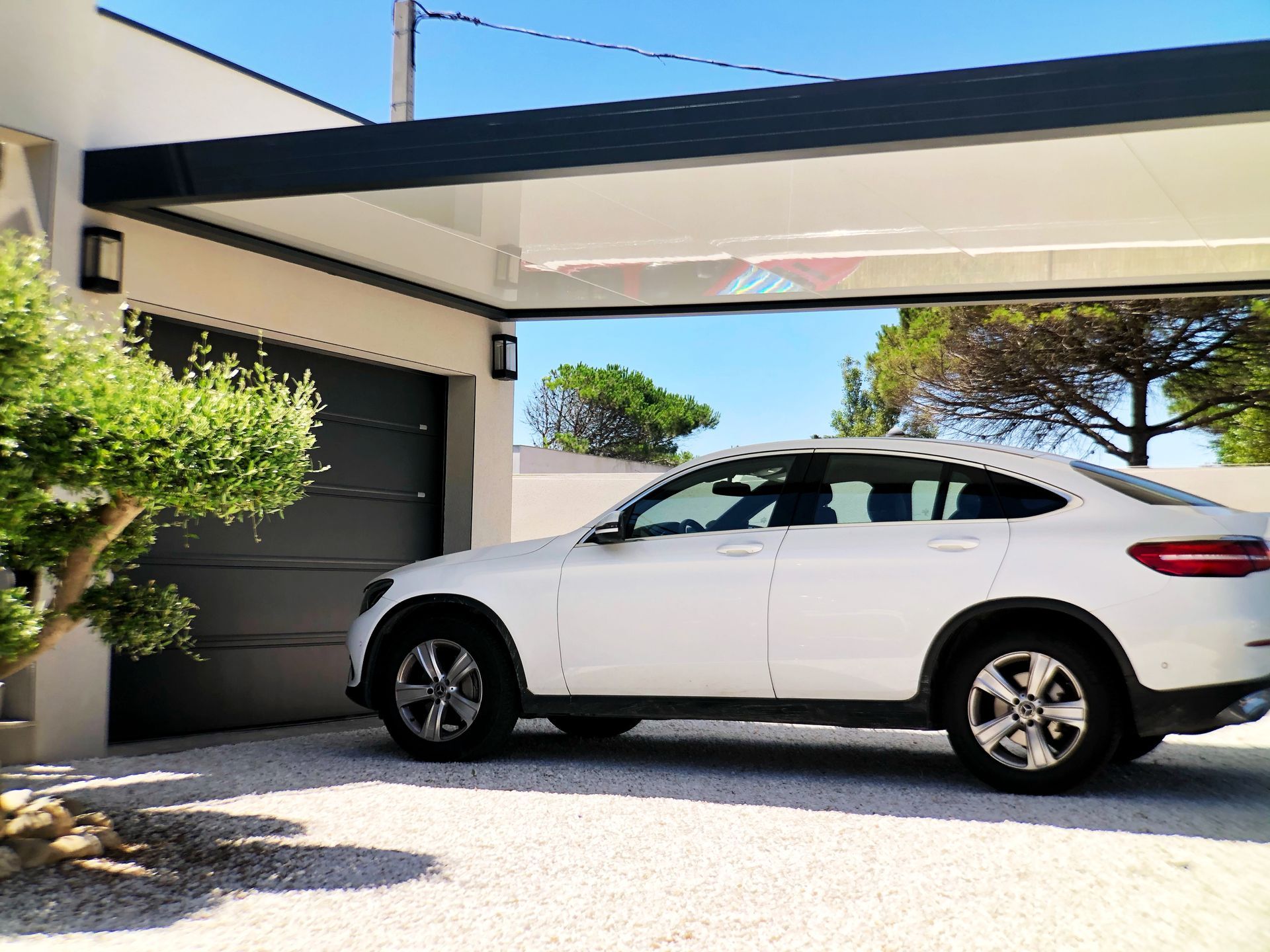 Voiture blanche garée sous un abri voiture devant un garage par une journée ensoleillée.