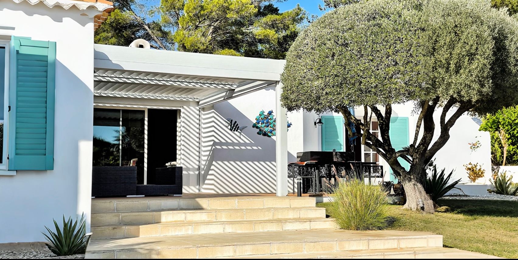 Maison blanche aux volets turquoise, olivier et marches menant à un porche couvert.