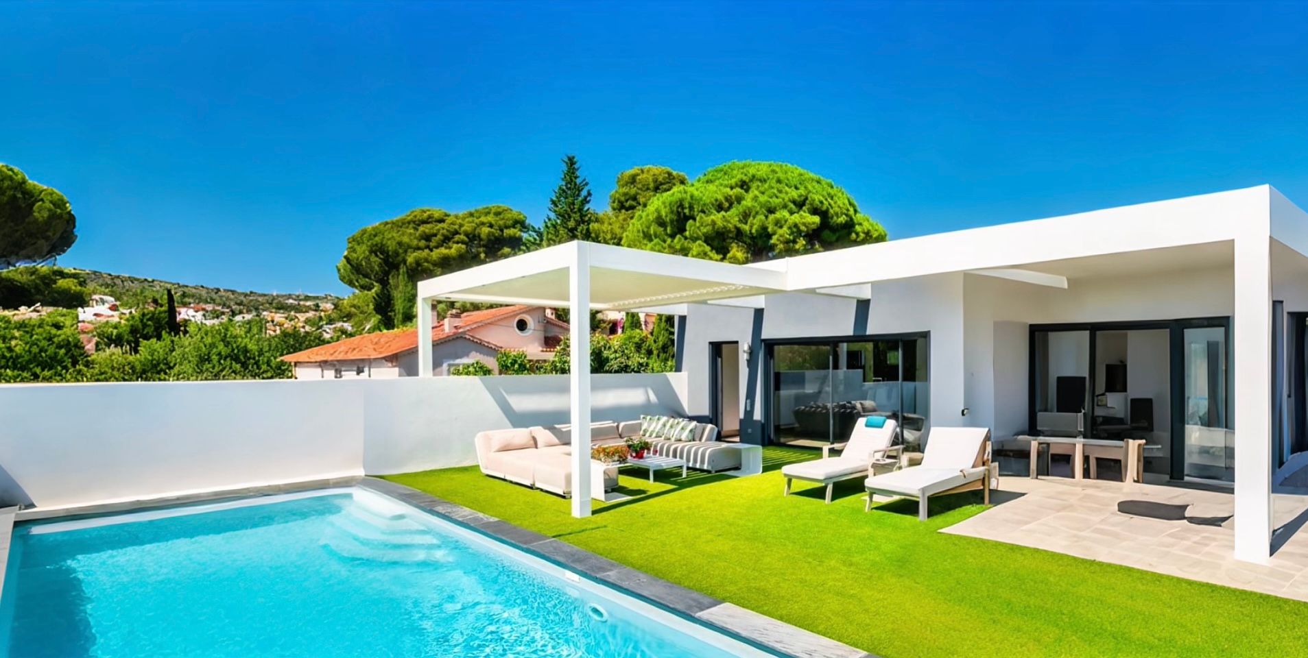 Une maison blanche moderne avec une piscine, une pelouse verte et un patio sous un ciel bleu.