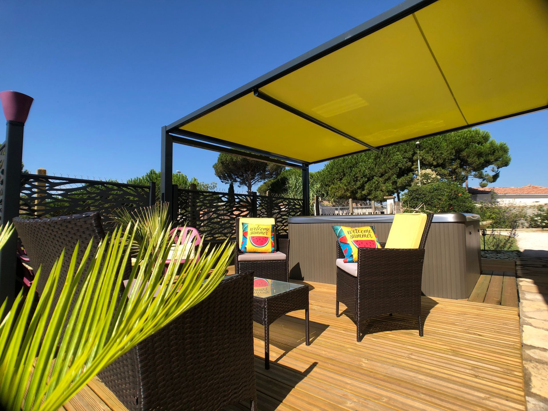 Terrasse extérieure avec jacuzzi et auvent jaune. Mobilier en osier, terrasse en bois, ciel bleu.