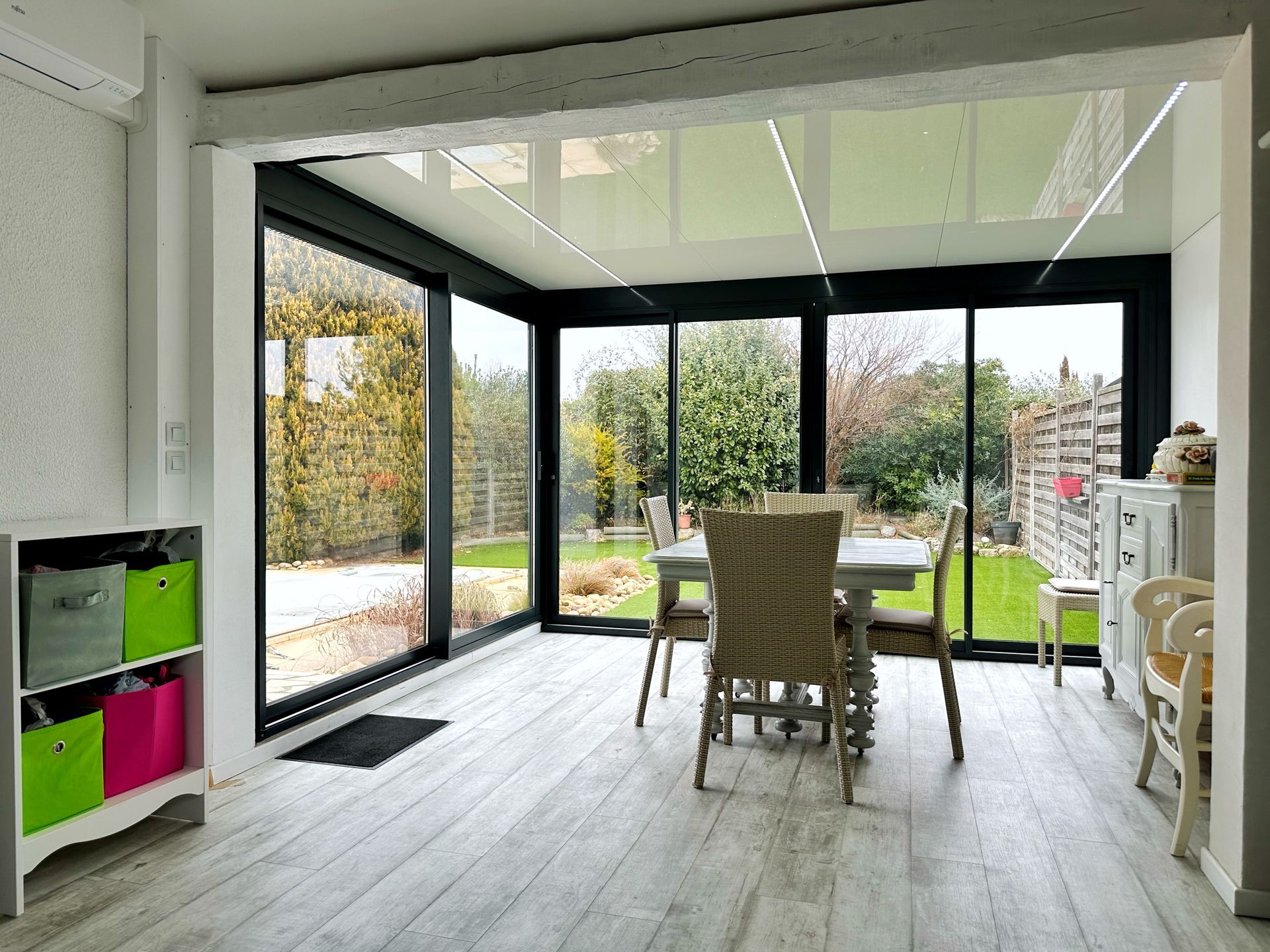 Véranda avec baies vitrées, table à manger et vue sur le jardin. Parquet clair, murs blancs et rangements colorés.