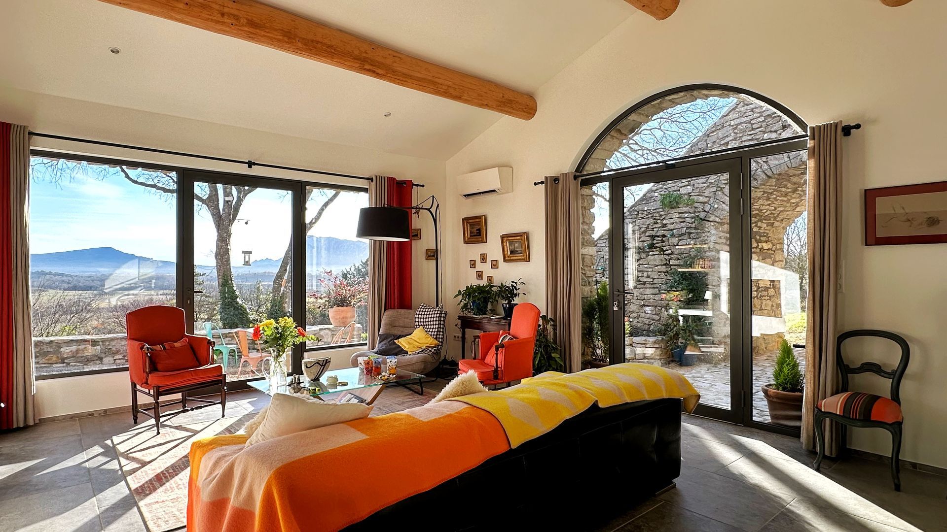 Salon avec portes vitrées cintrées donnant sur une terrasse et un paysage. Touches orange et rouge.
