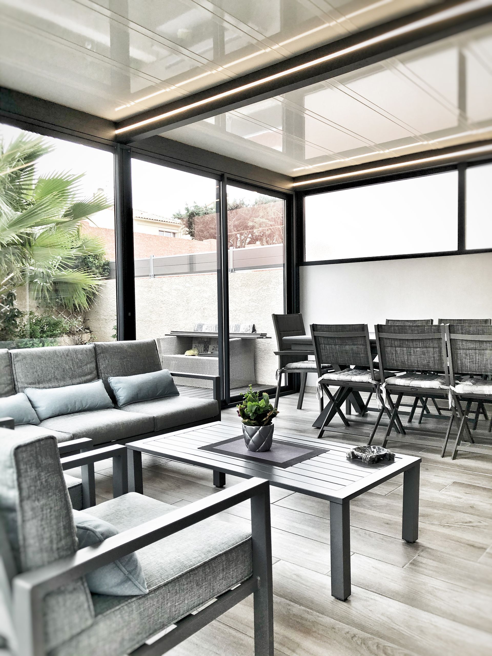 Chambre extérieure avec mobilier gris, murs en verre et vue sur un jardin.