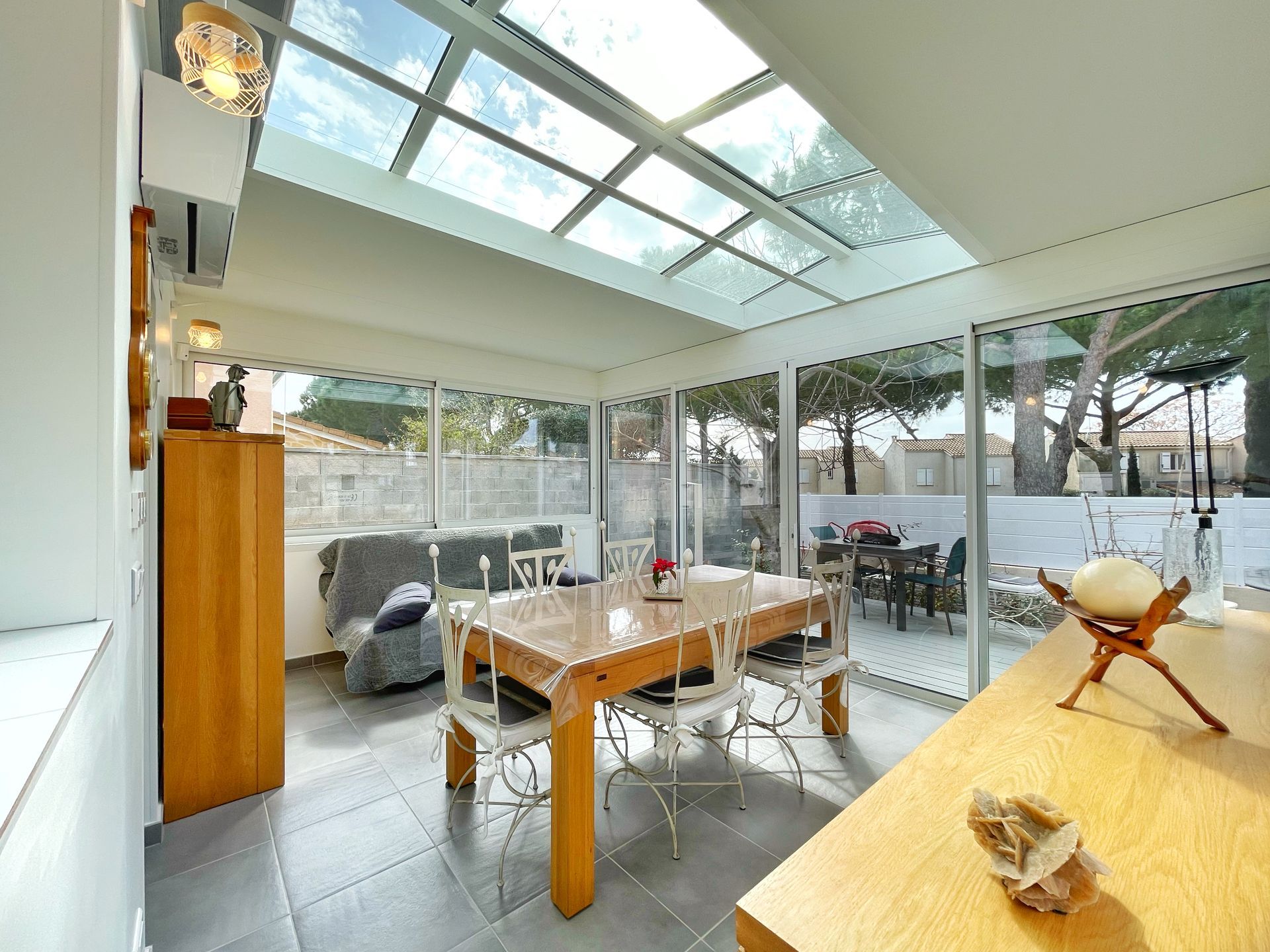 Véranda avec toit en verre, table à manger, chaises et vue sur les arbres extérieurs.