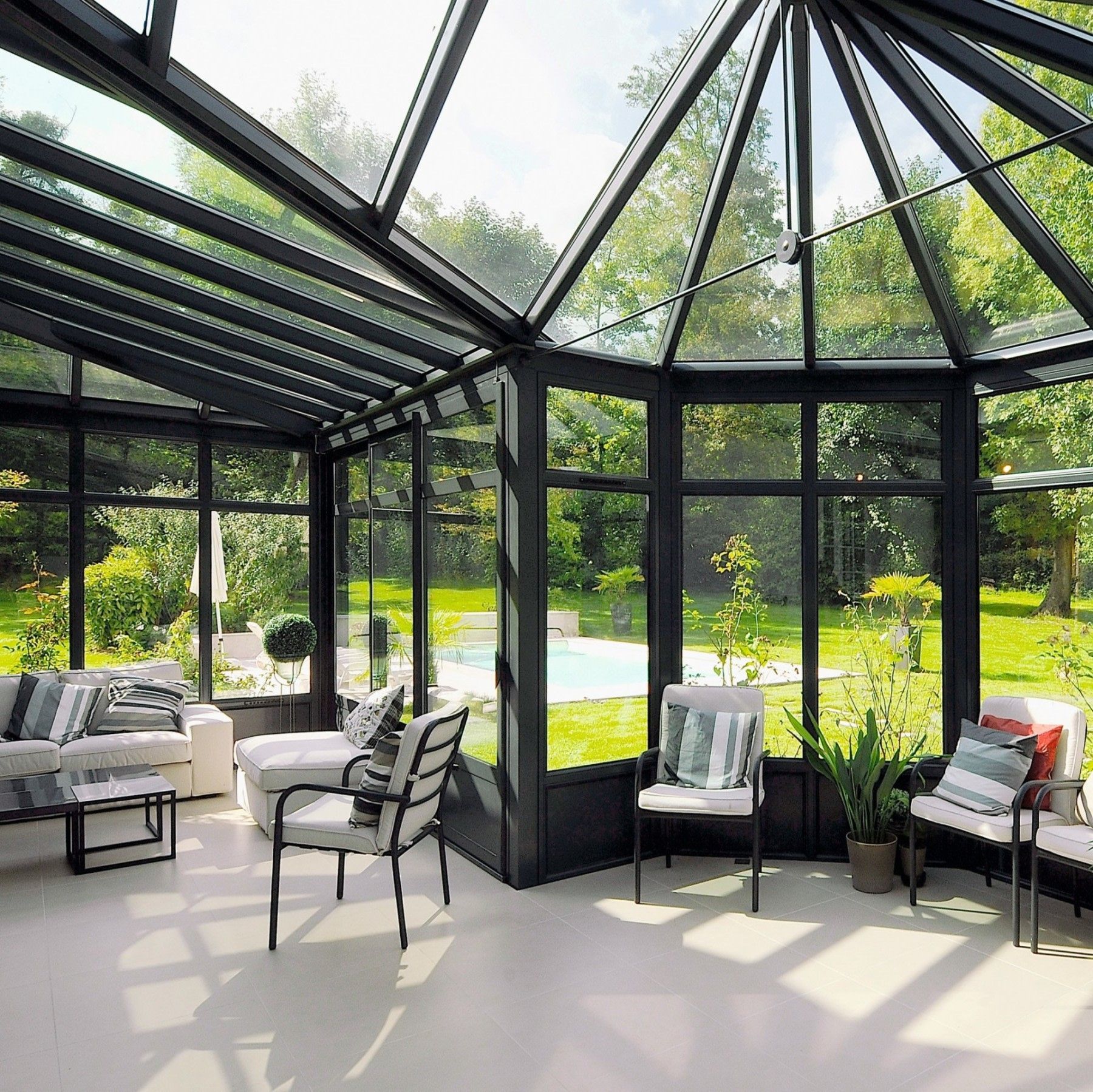 Véranda avec murs et toit en verre à cadre noir, mobilier blanc et vue sur une cour arrière avec piscine et pelouse verte.
