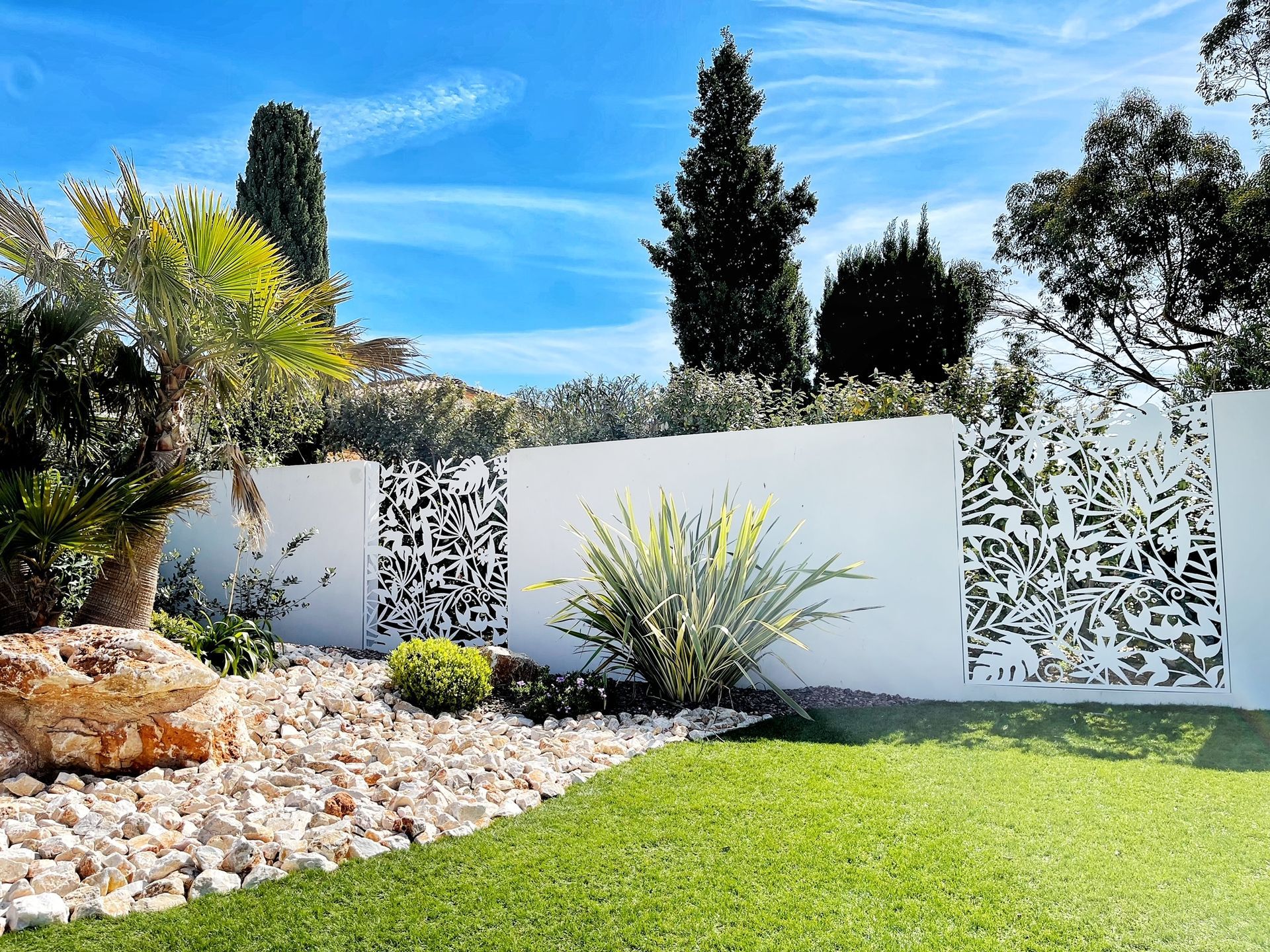 Clôture blanche avec découpes décoratives, jardin luxuriant, herbe verte, ciel bleu.
