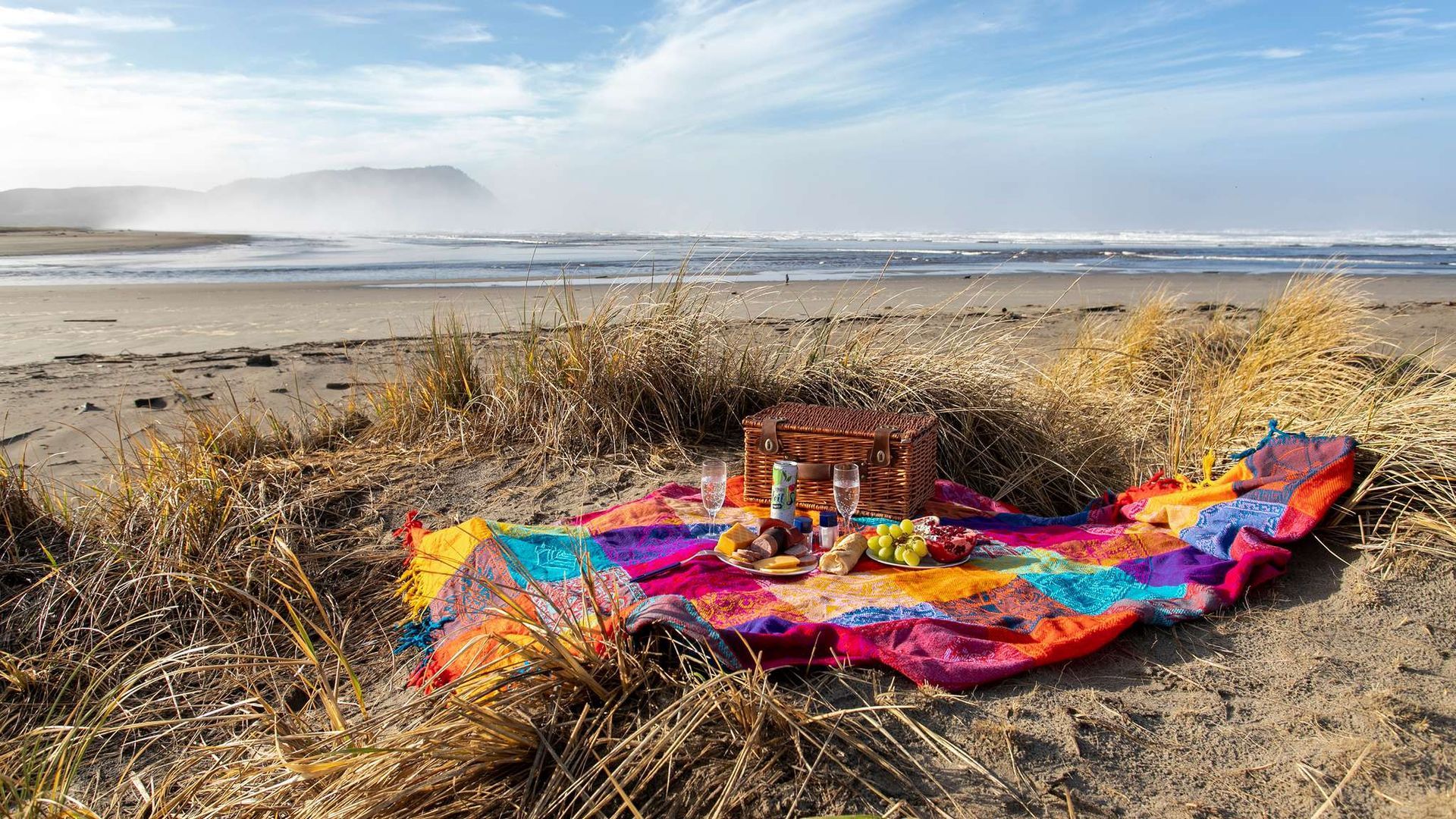 Picnic on a beach; colorful blanket, wicker basket, ocean waves in background, sunny day.