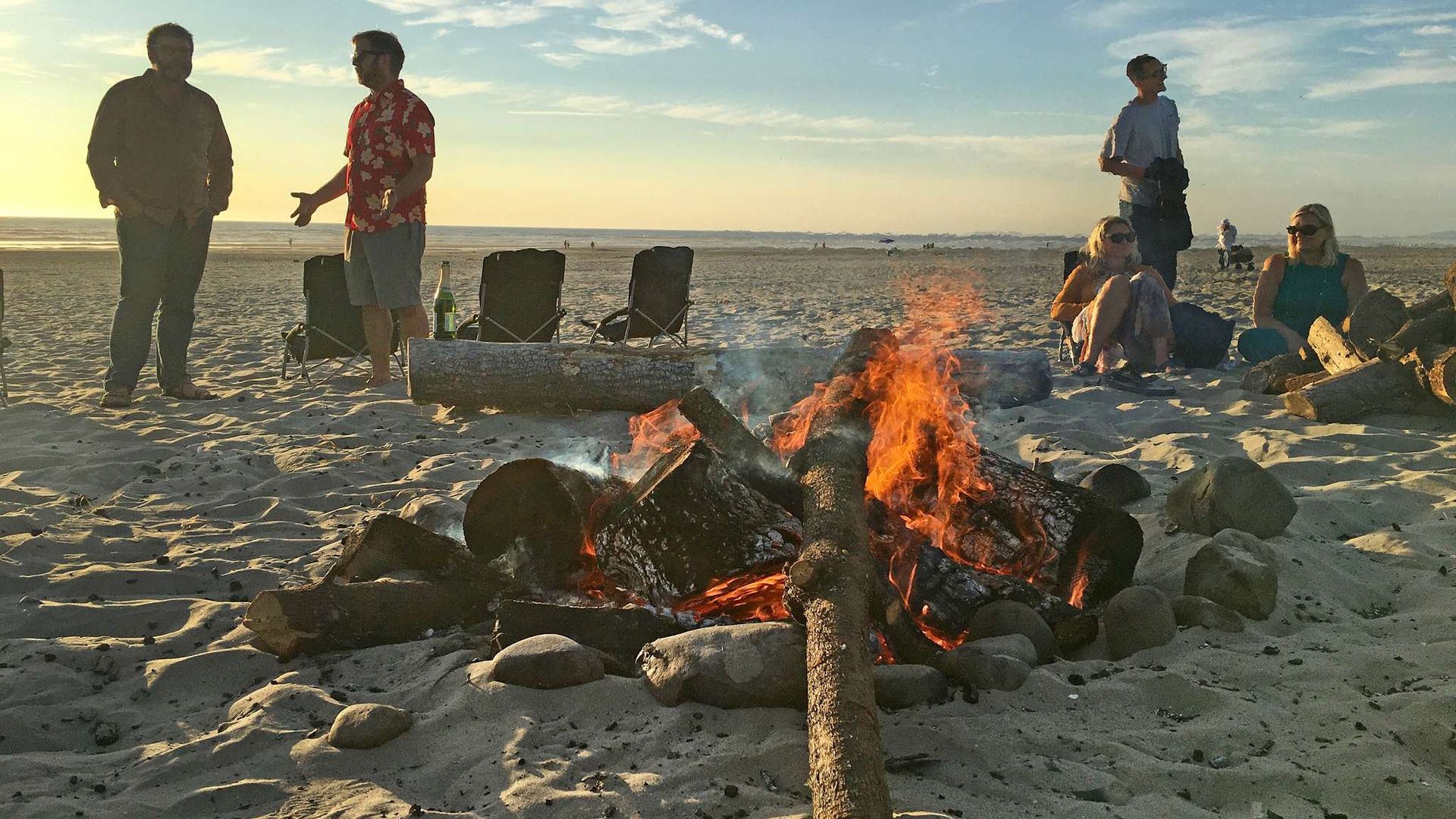 Campfire on a beach at sunset with a small group of people gathered around it.