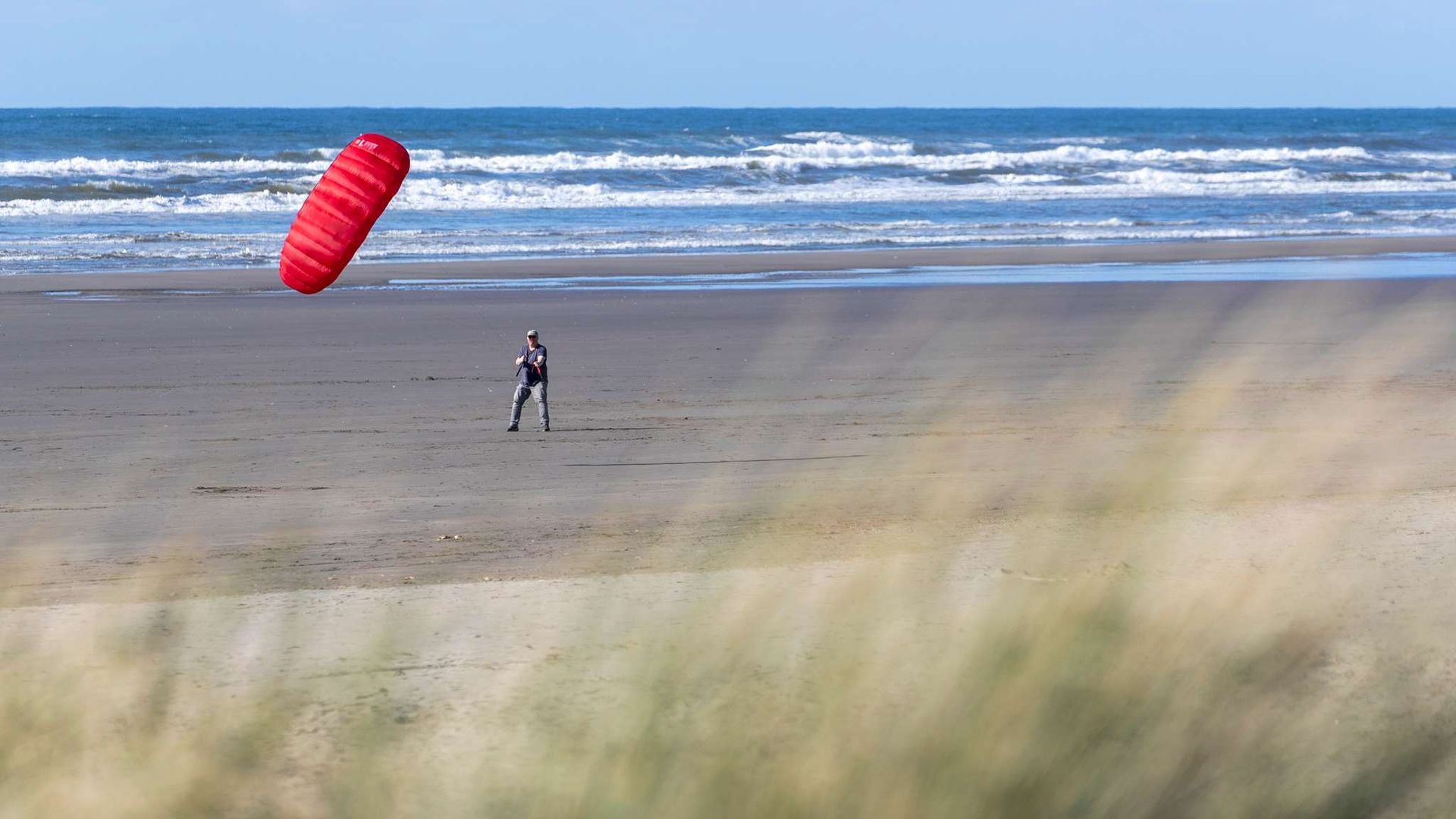 Person flying a red kite on a sandy beach. Ocean and blue sky in background.