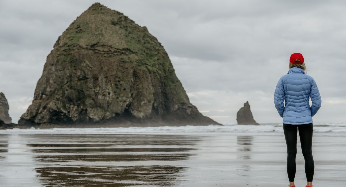 A woman in a blue puffer jacket and red cap stands on a wet sandy beach in Oregon, gazing towards a