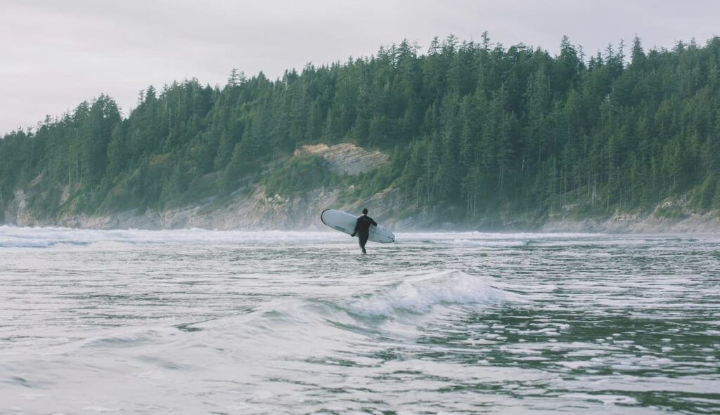 A man carrying a surfboard in the ocean with green forests in the background on the Oregon coast