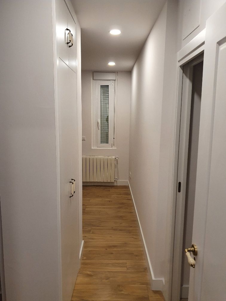 Un largo pasillo con pisos de madera y paredes blancas que conduce a una ventana.