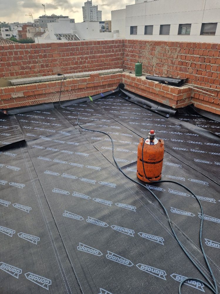 Un cilindro de gas está colocado encima de un techo con una pared de ladrillos al fondo.