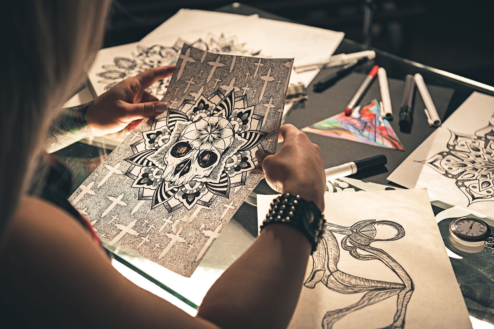 Persona sosteniendo una obra de arte detallada en blanco y negro, rodeada de dibujos sobre una mesa de cristal.