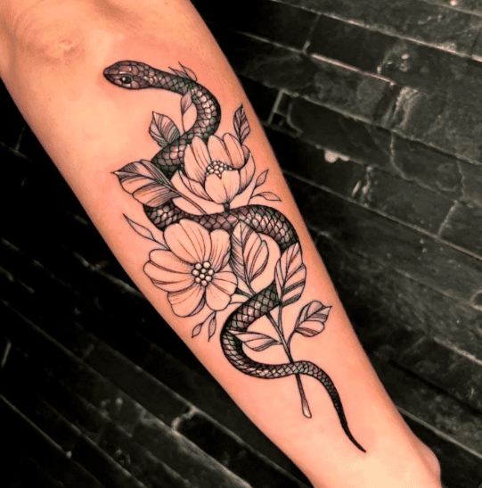 Tatuaje con tinta negra de una serpiente enroscada alrededor de flores y hojas blancas en el antebrazo.
