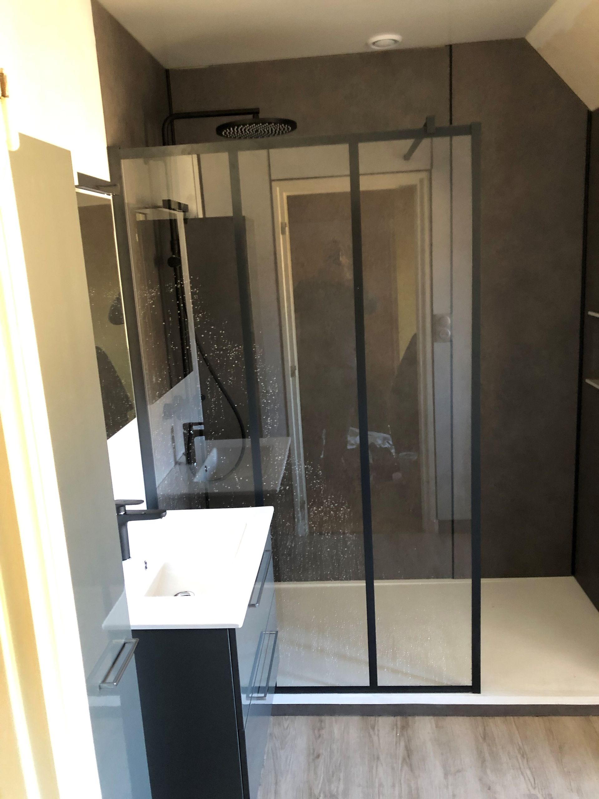 Salle de bains moderne avec douche en verre, murs gris foncé et lavabo blanc.