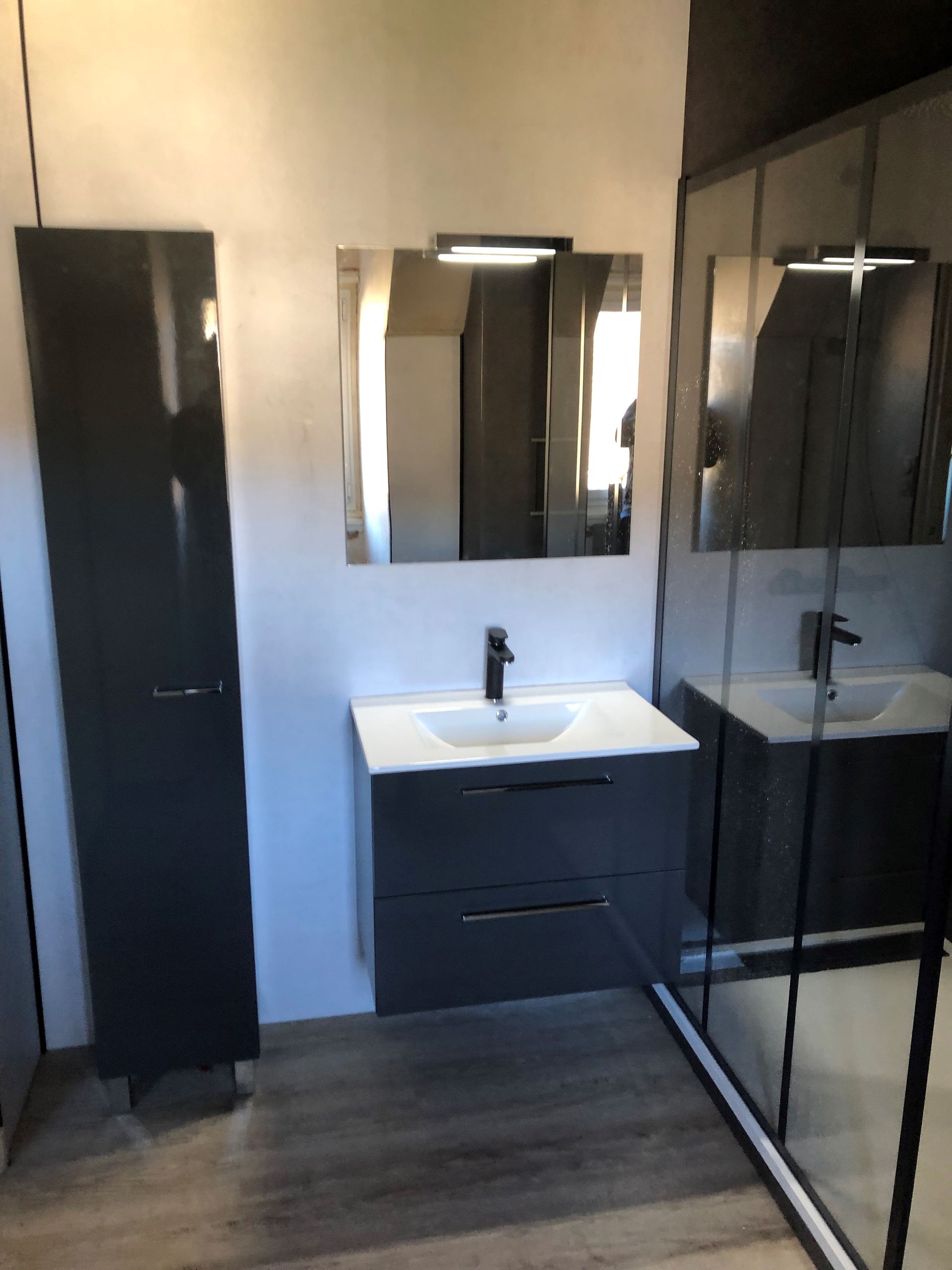Salle de bains moderne avec meuble-lavabo gris foncé, miroir et armoire haute. Douche avec porte vitrée visible.