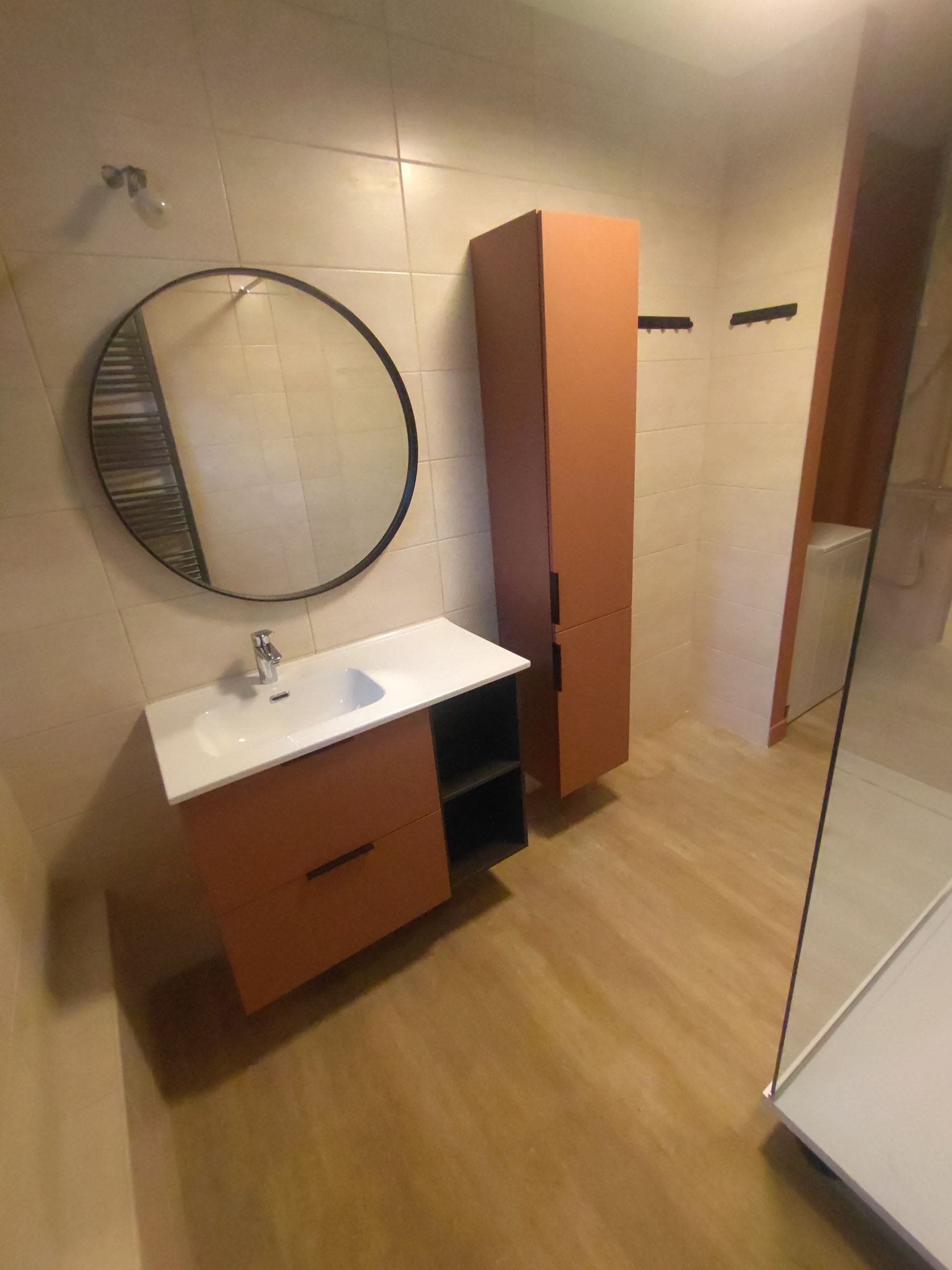 Salle de bains avec meubles orange, miroir rond et douche avec porte en verre.