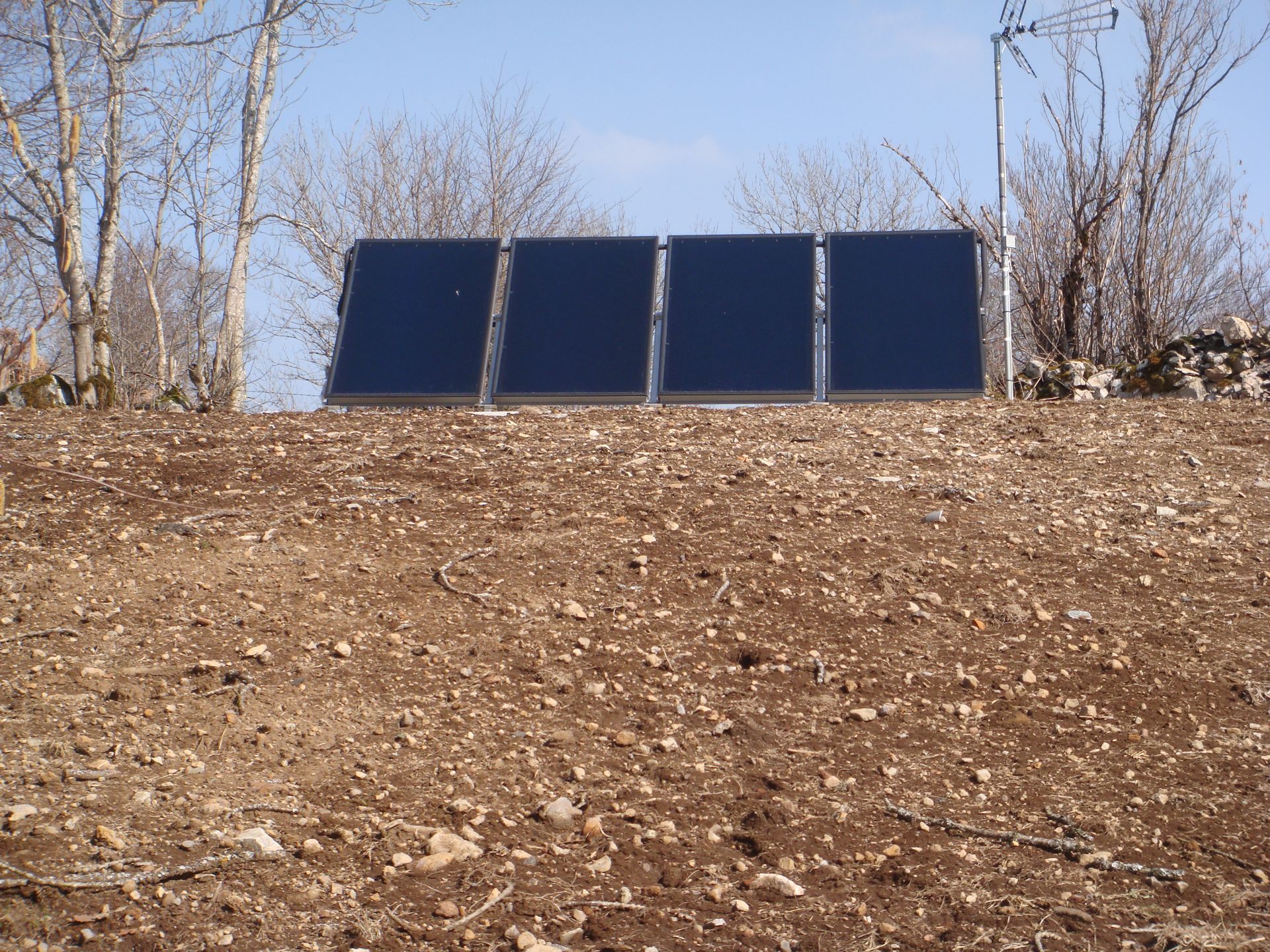 Quatre panneaux solaires sur un flanc de colline brun se détachant sur un ciel bleu, avec des arbres dénudés et une antenne radio en arrière-plan.