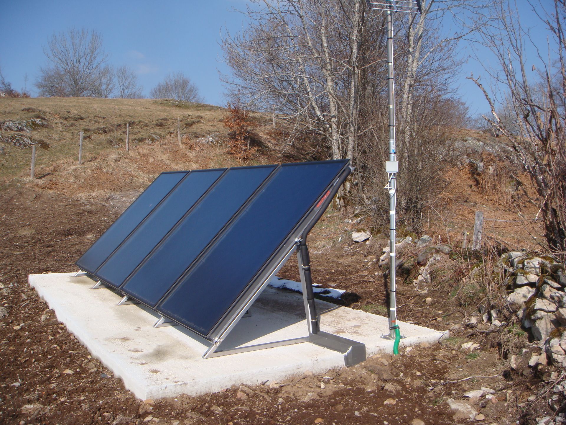Panneaux solaires installés sur une base en béton à l'extérieur, avec un tuyau blanc et des arbres en arrière-plan.