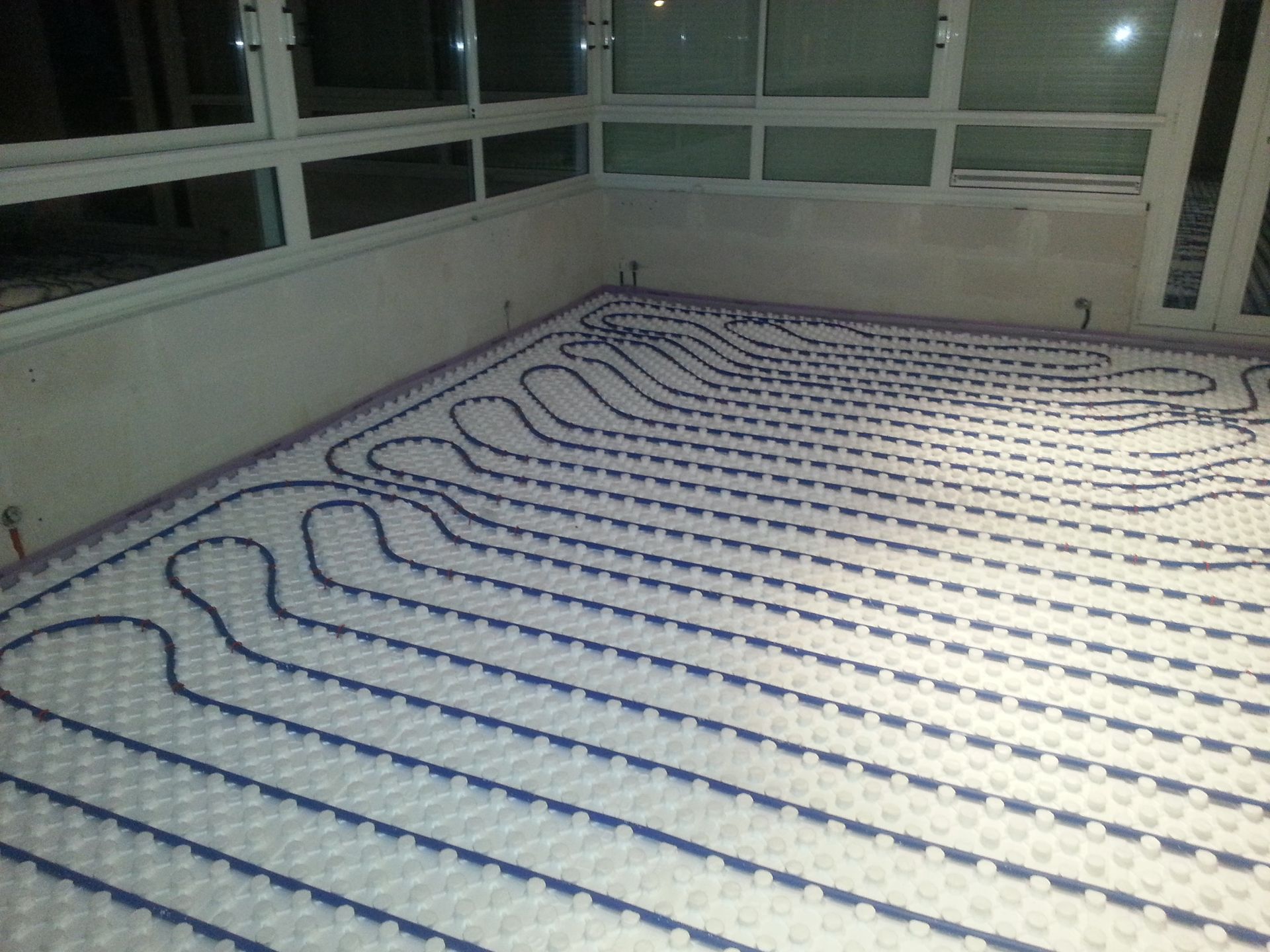 Des tuyaux bleus disposés en grille sur une sous-couche blanche, pour un chauffage au sol radiant, dans une pièce avec fenêtres.