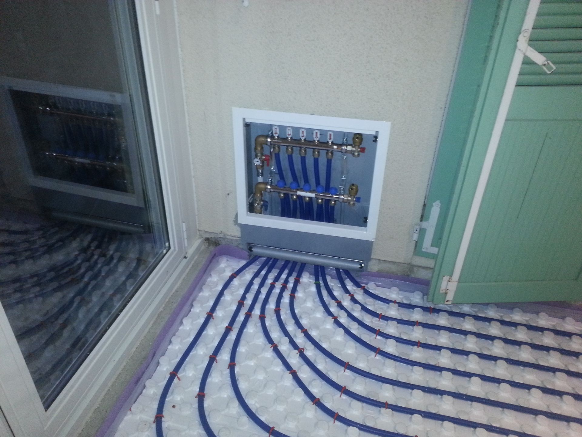 Tuyaux bleus pour chauffage au sol raccordés à un collecteur de distribution, près d'une fenêtre et d'une porte.