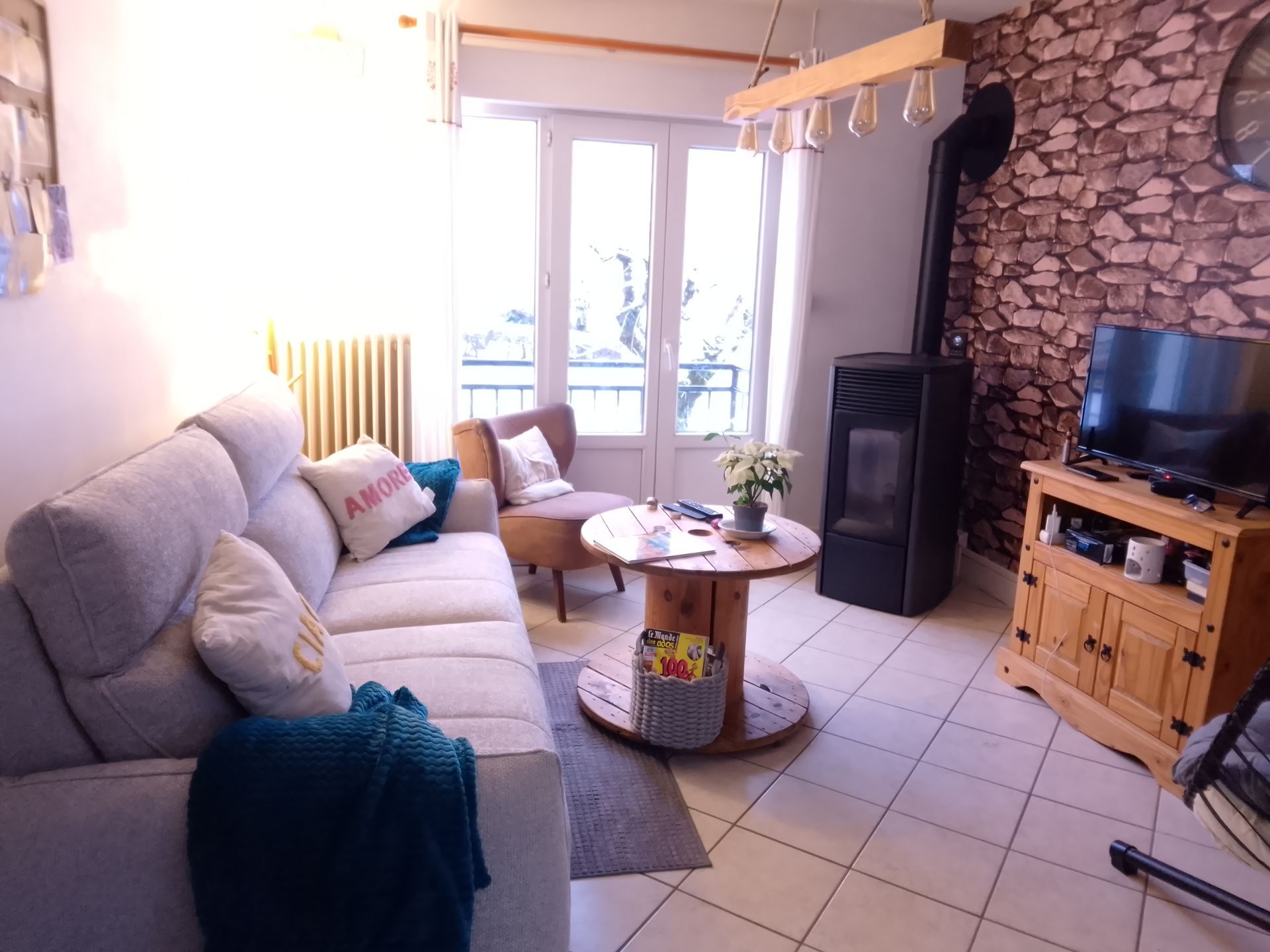 Salon confortable avec canapé, fauteuil, télévision et poêle à bois, avec un mur de briques et une fenêtre.
