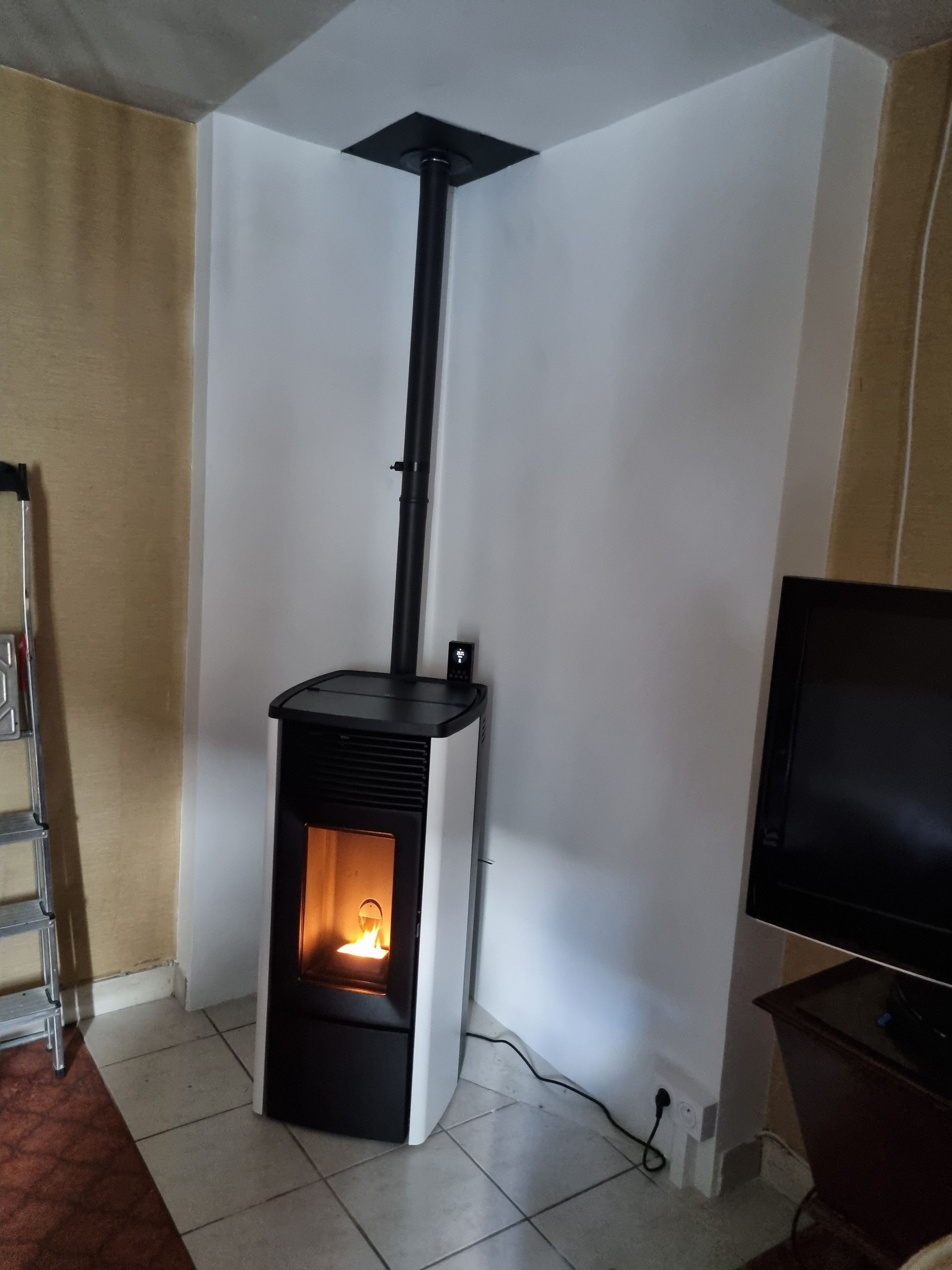 Poêle à granulés blanc et noir avec cheminée noire, installé en angle, avec un feu allumé visible.