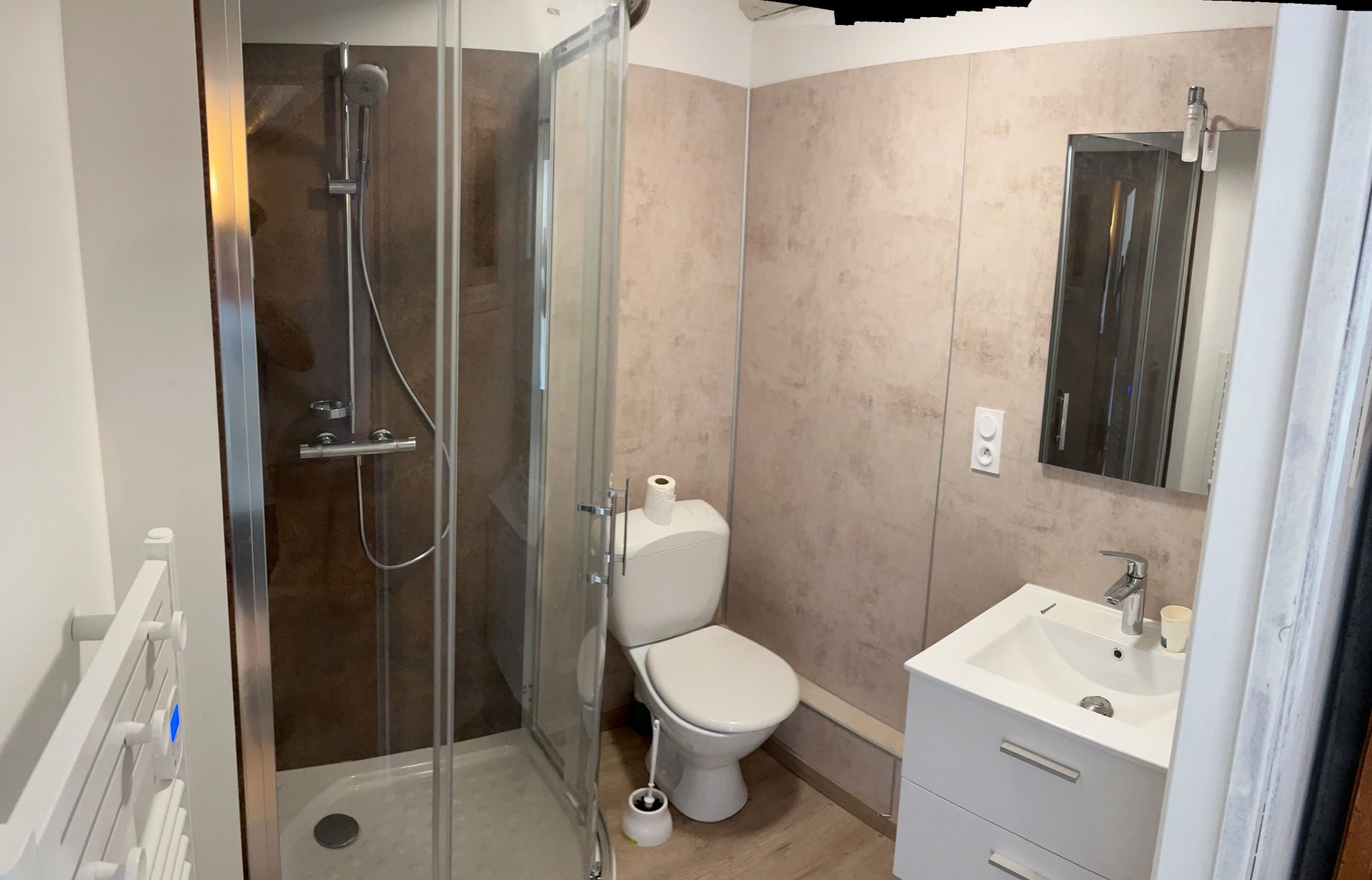 Salle de bains avec douche, toilettes, lavabo et miroir. Murs de douche marron, murs beiges et robinetterie blanche.