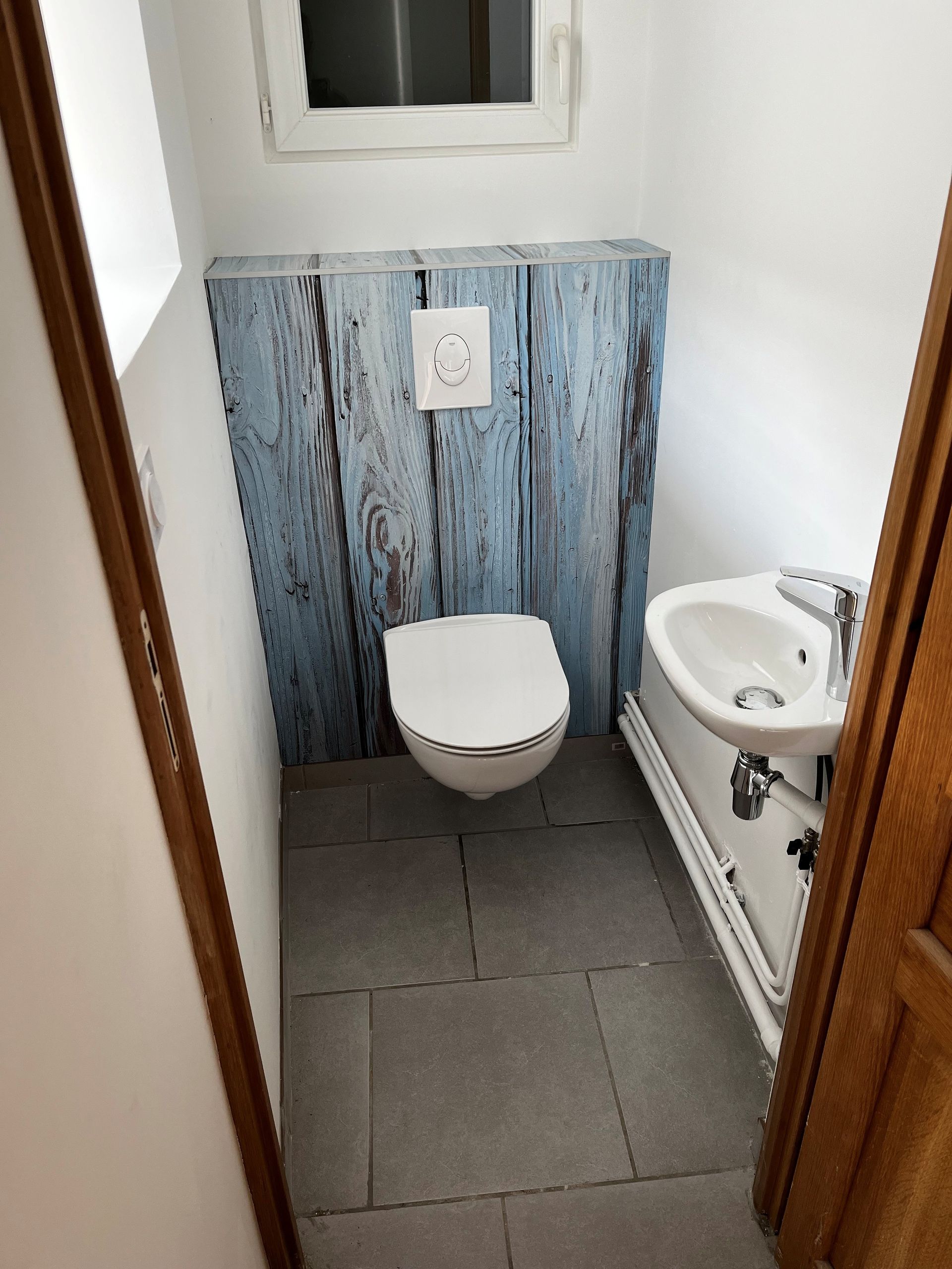 Petite salle de bains avec toilettes, lavabo et sol carrelé gris ; mur en bois bleu derrière les toilettes.