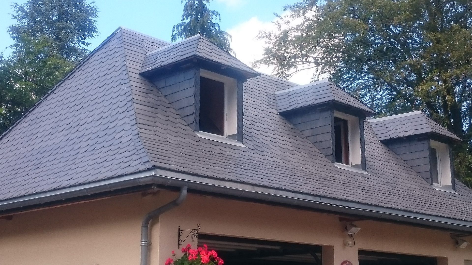 Toit en ardoise avec trois lucarnes sur un bâtiment de couleur claire, arbres en arrière-plan.