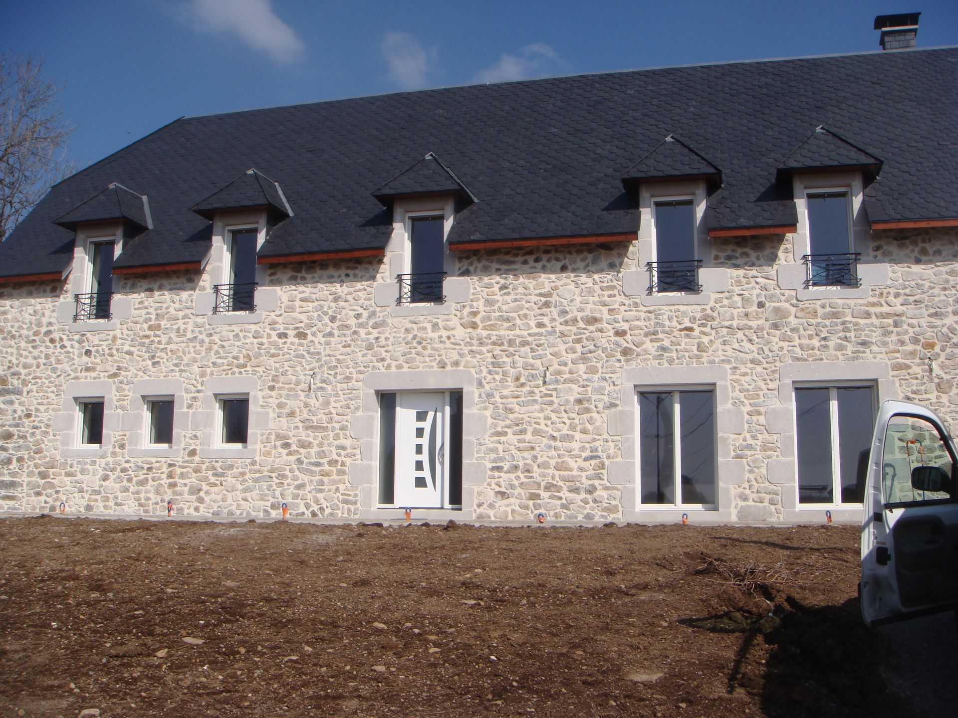 Maison en pierre avec toit d'ardoise, lucarnes et fenêtres à cadre blanc, par une journée ensoleillée.
