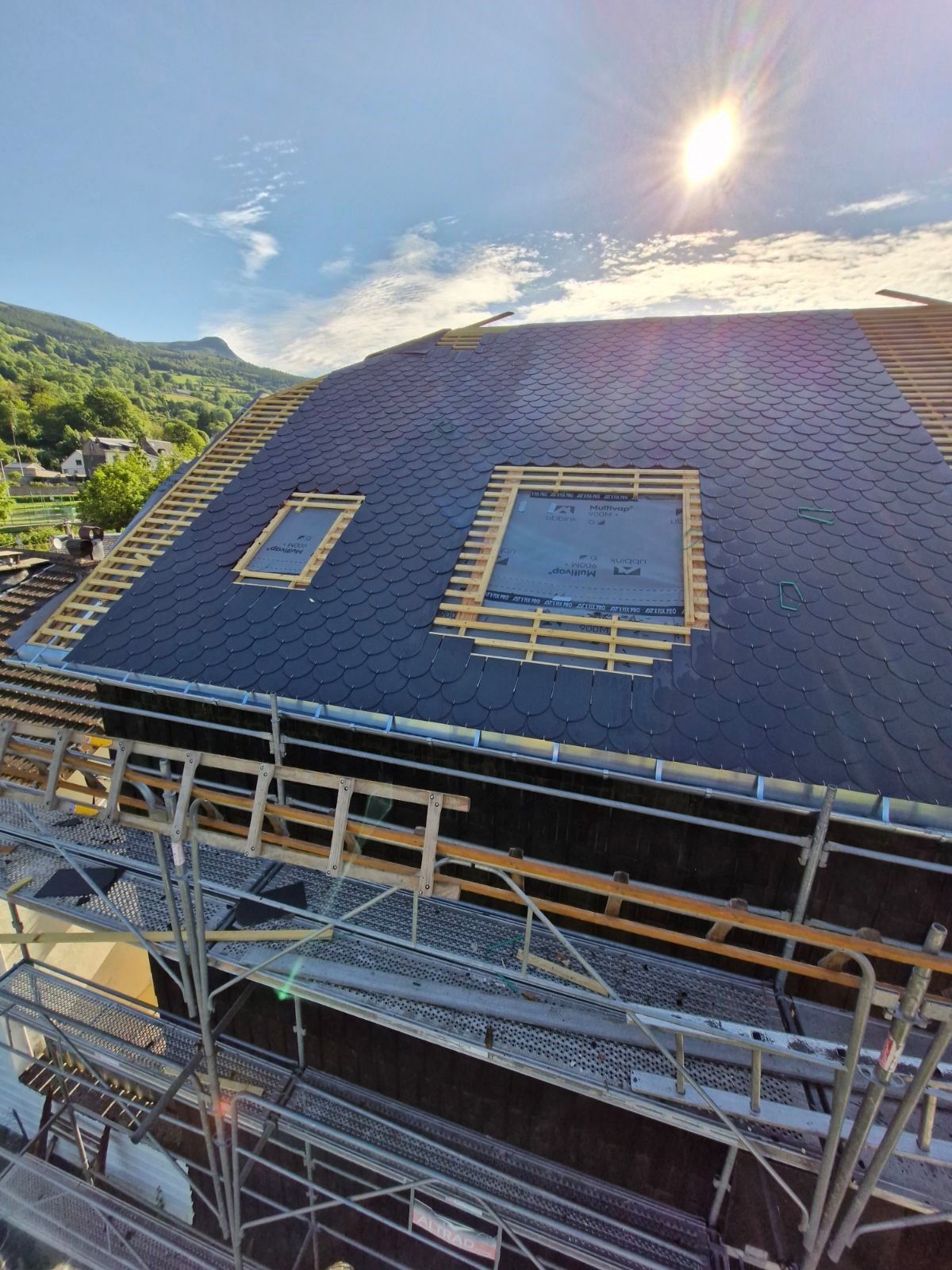 Toiture en construction avec bardeaux noirs, deux puits de lumière, échafaudages et montagnes en arrière-plan sous un soleil éclatant.